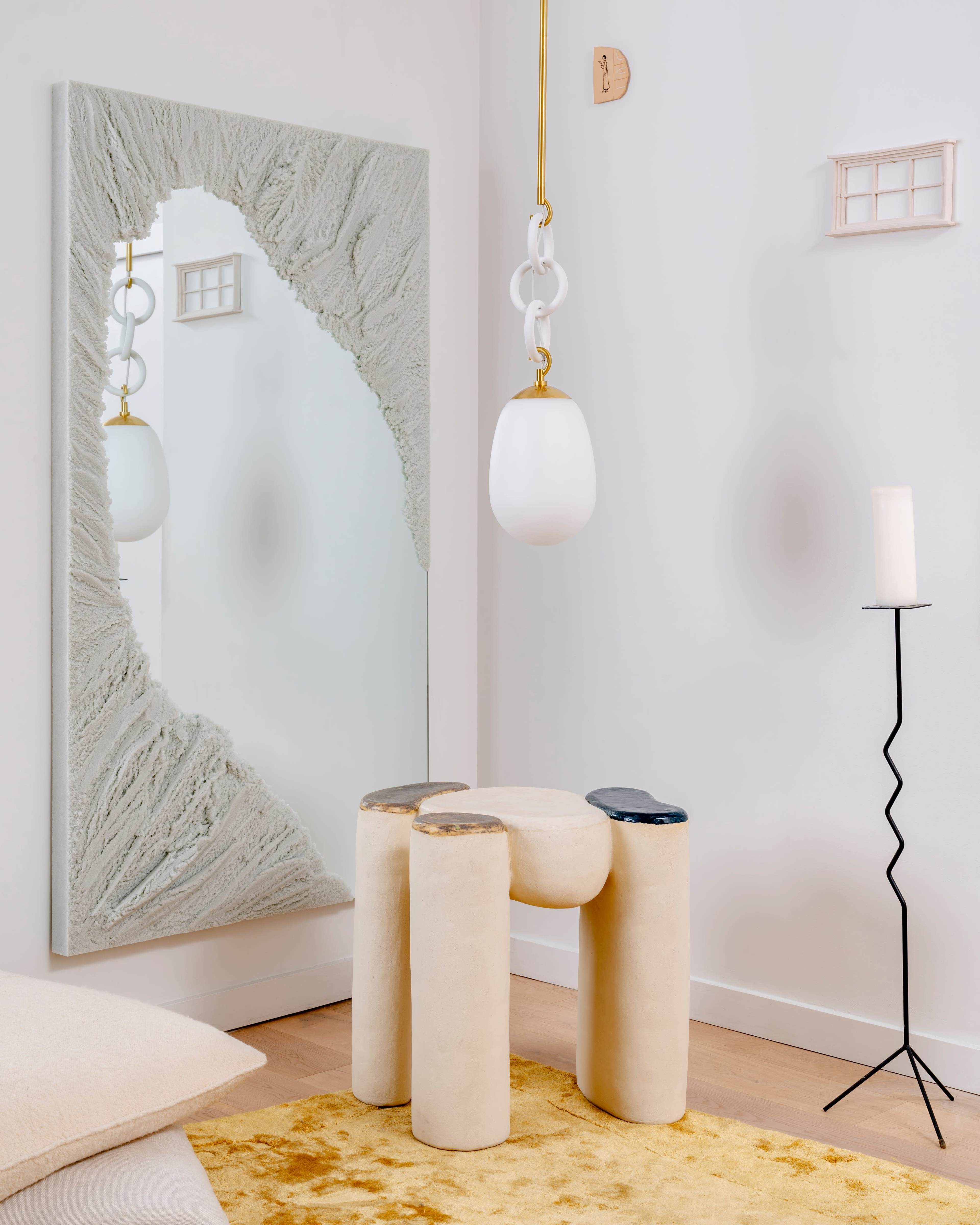 A modern interior with a textured mirror, a pendant light, a sculptural stool, a candle holder, and a yellow rug.