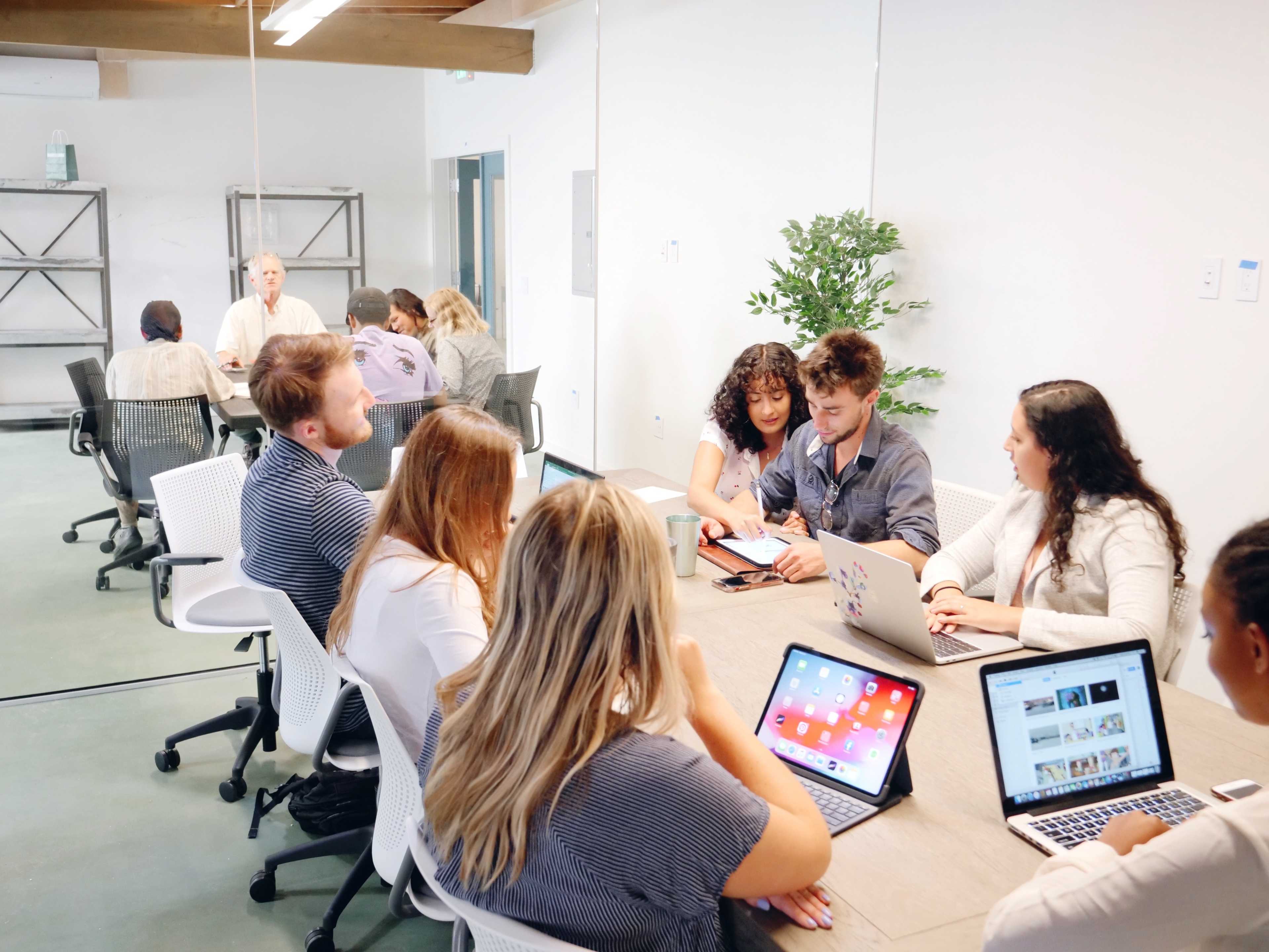 A group of people is seated around a large table in a conference room, engaging in discussions and working on laptops.