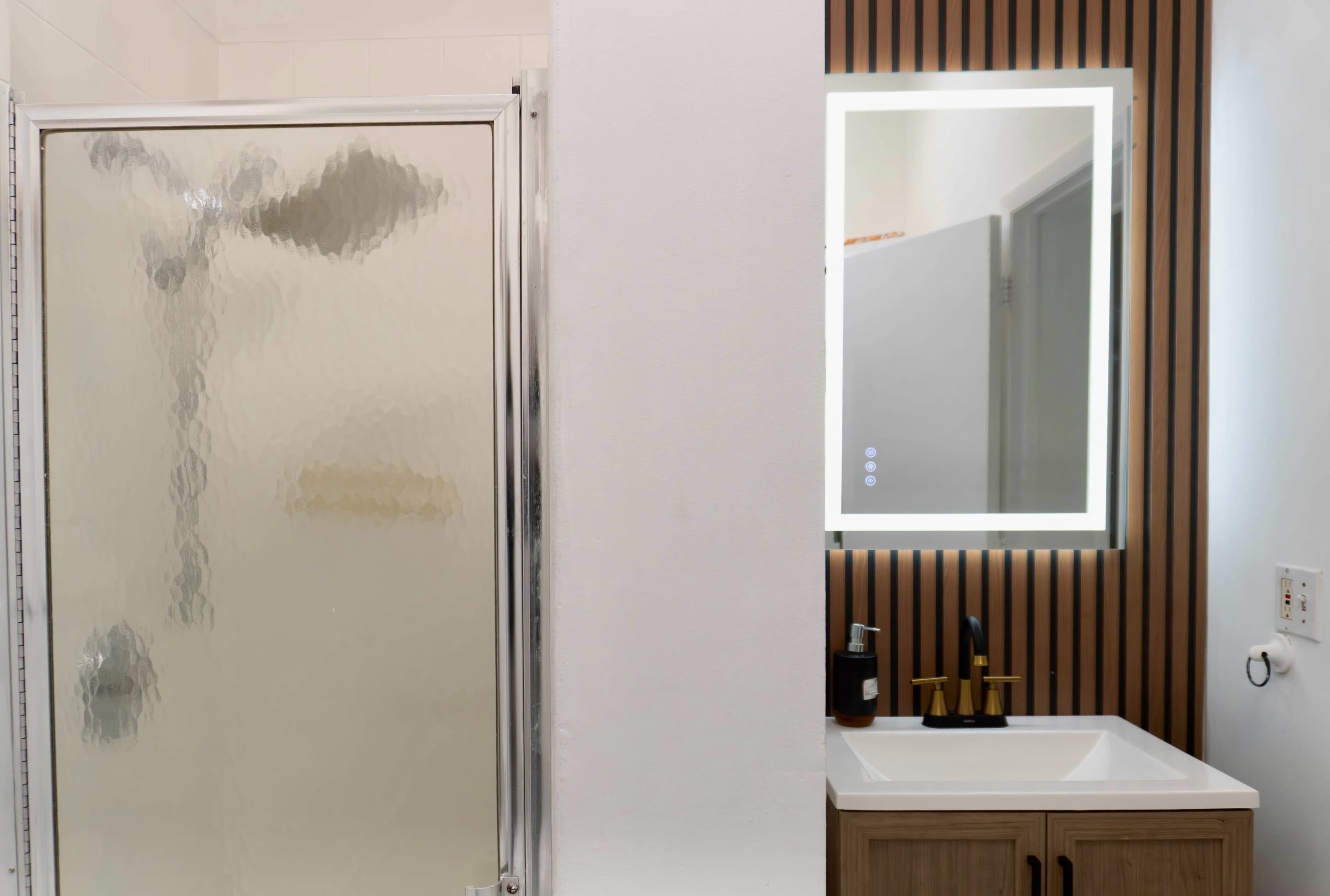 The image shows a bathroom featuring a glass shower stall on the left and a vanity with a mirror and faucet on the right, separated by a wall.