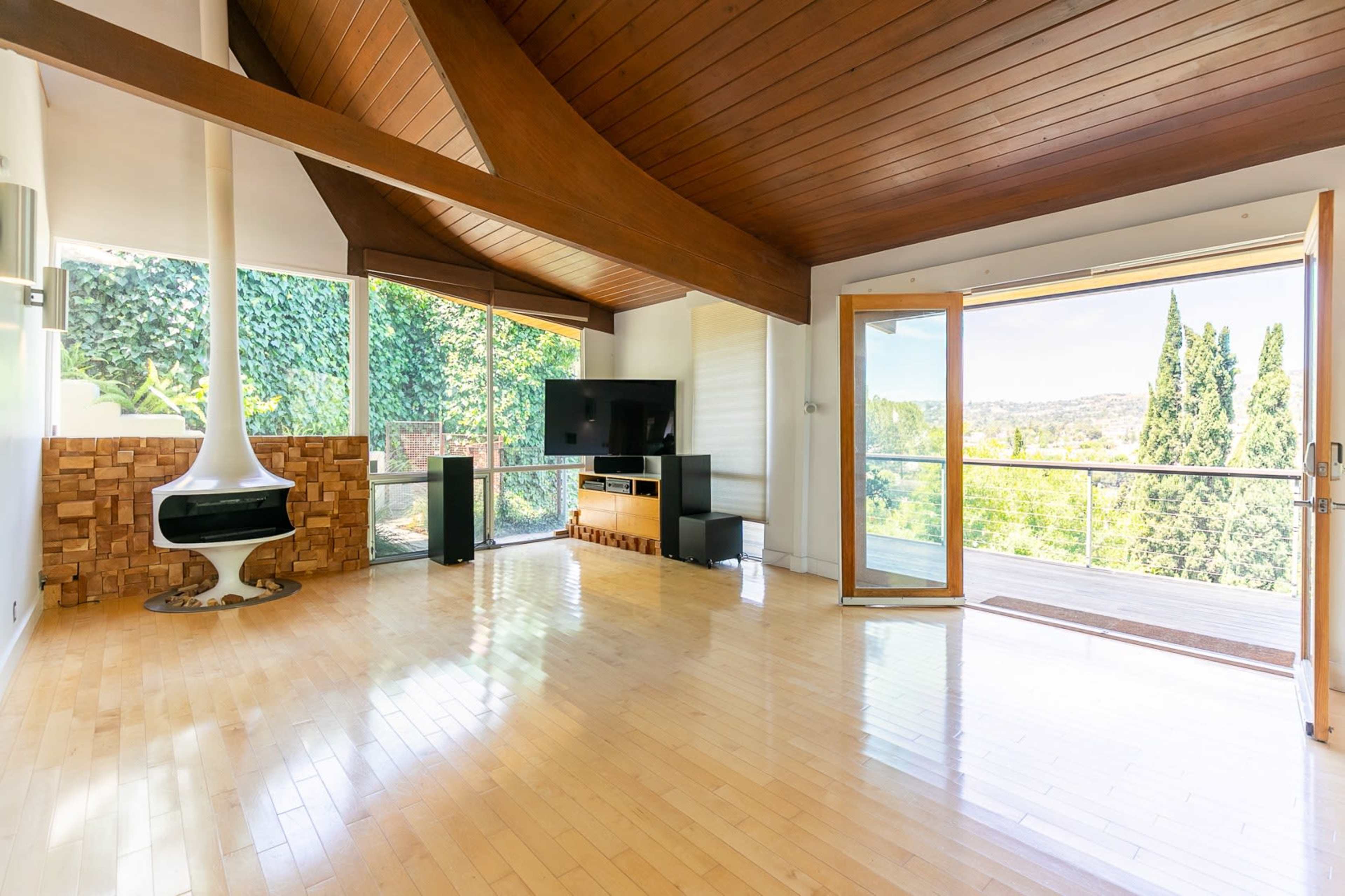 The image shows a modern living room with large windows, wooden beams, and a white fireplace, overlooking a lush outdoor view.