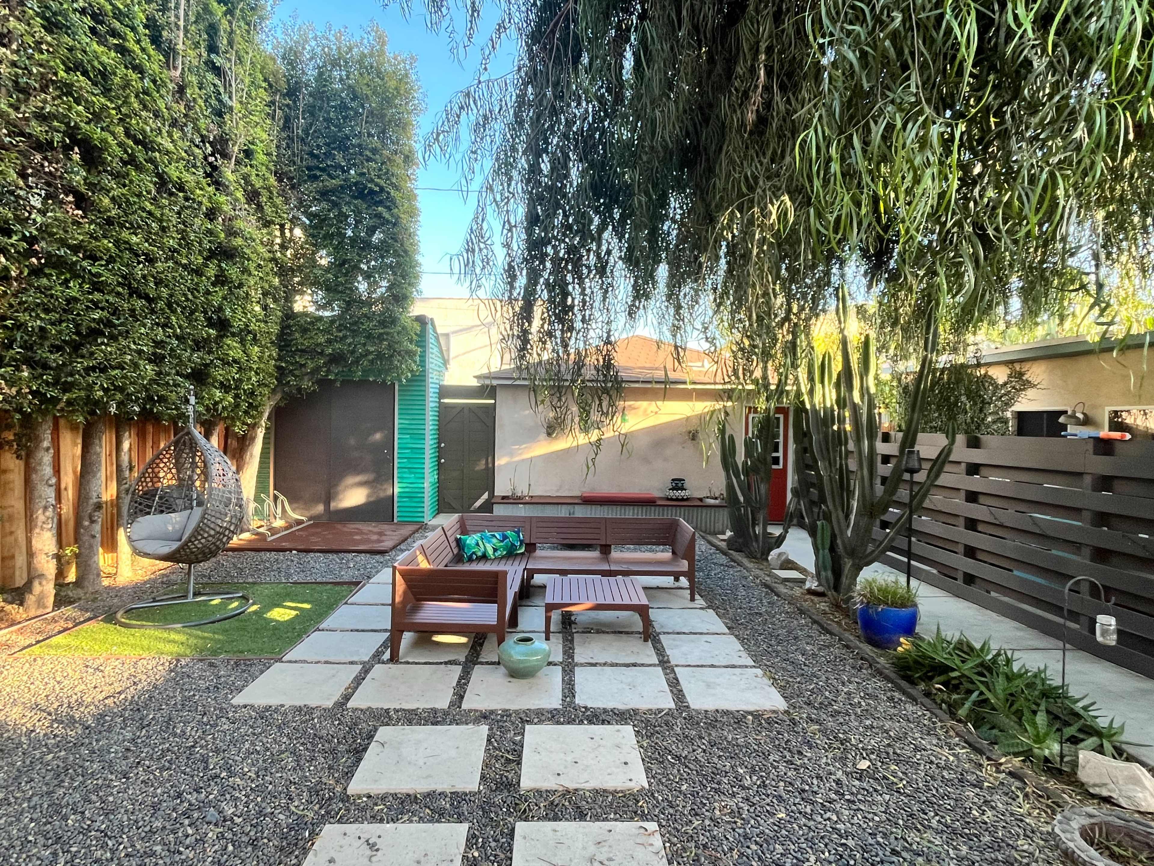 The image shows a landscaped outdoor area featuring stone pavers, wooden furniture, a hanging chair, and various plants.