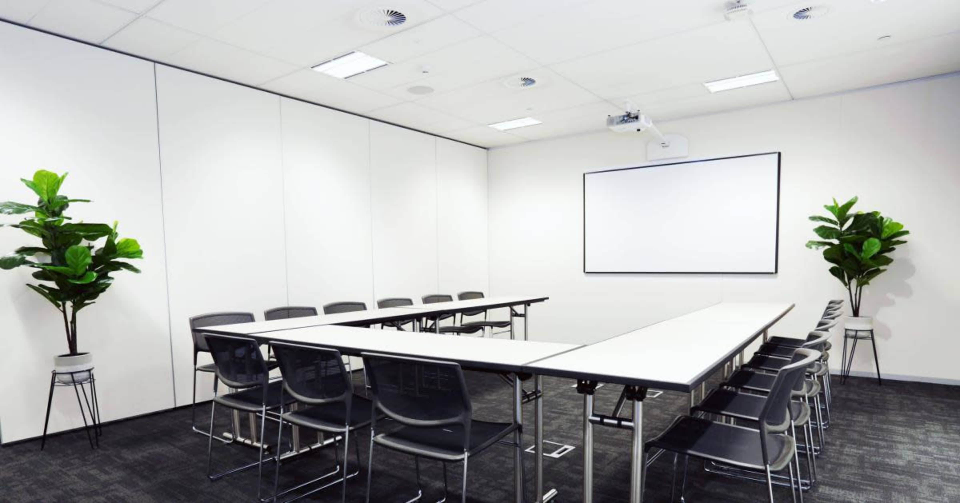 A modern conference room with a long table surrounded by black chairs, two potted plants in the corners, and a whiteboard on the wall.
