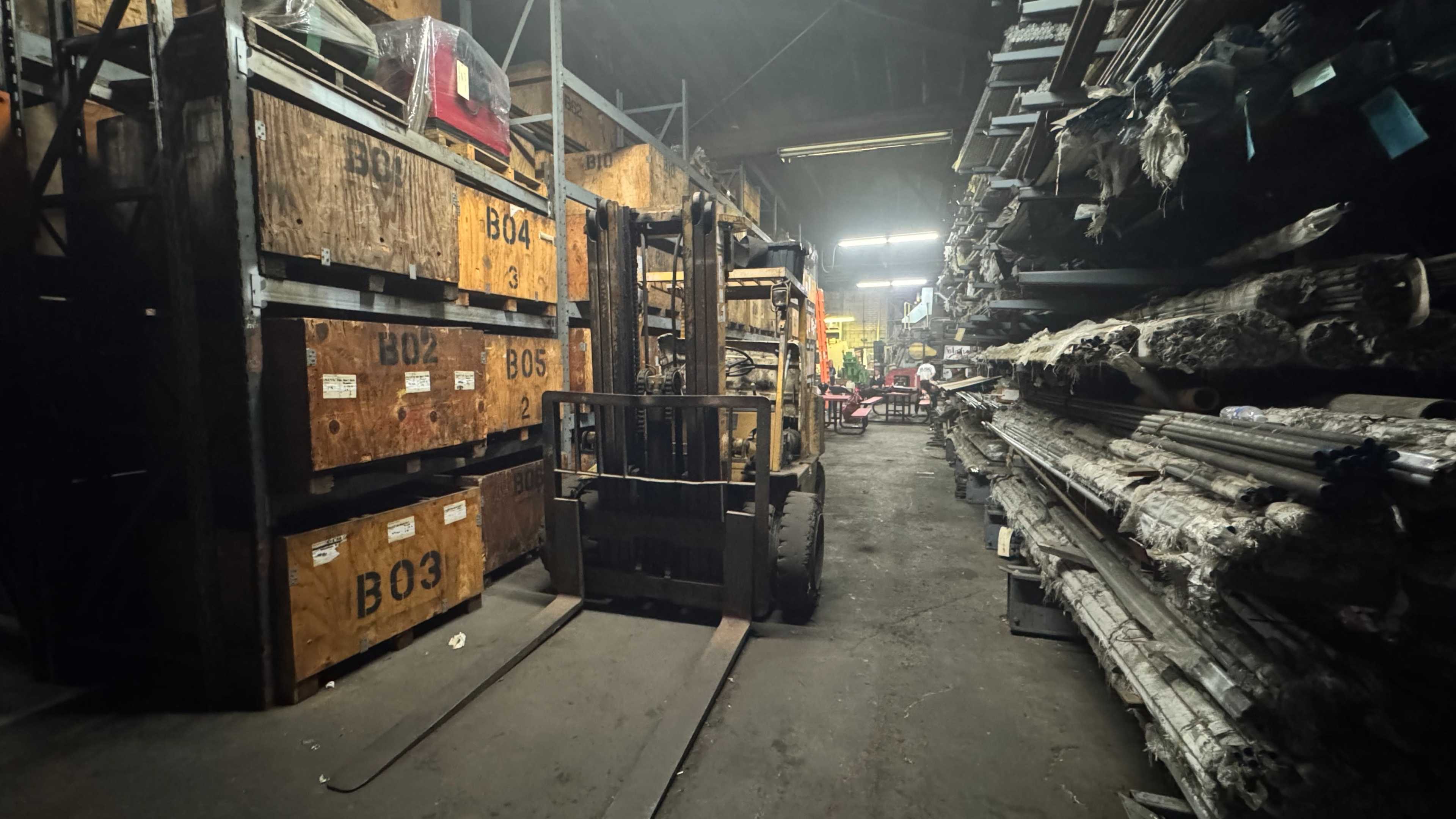 The image shows a warehouse aisle with a forklift positioned between wooden storage crates labeled "B02," "B03," and other items stacked on shelves.