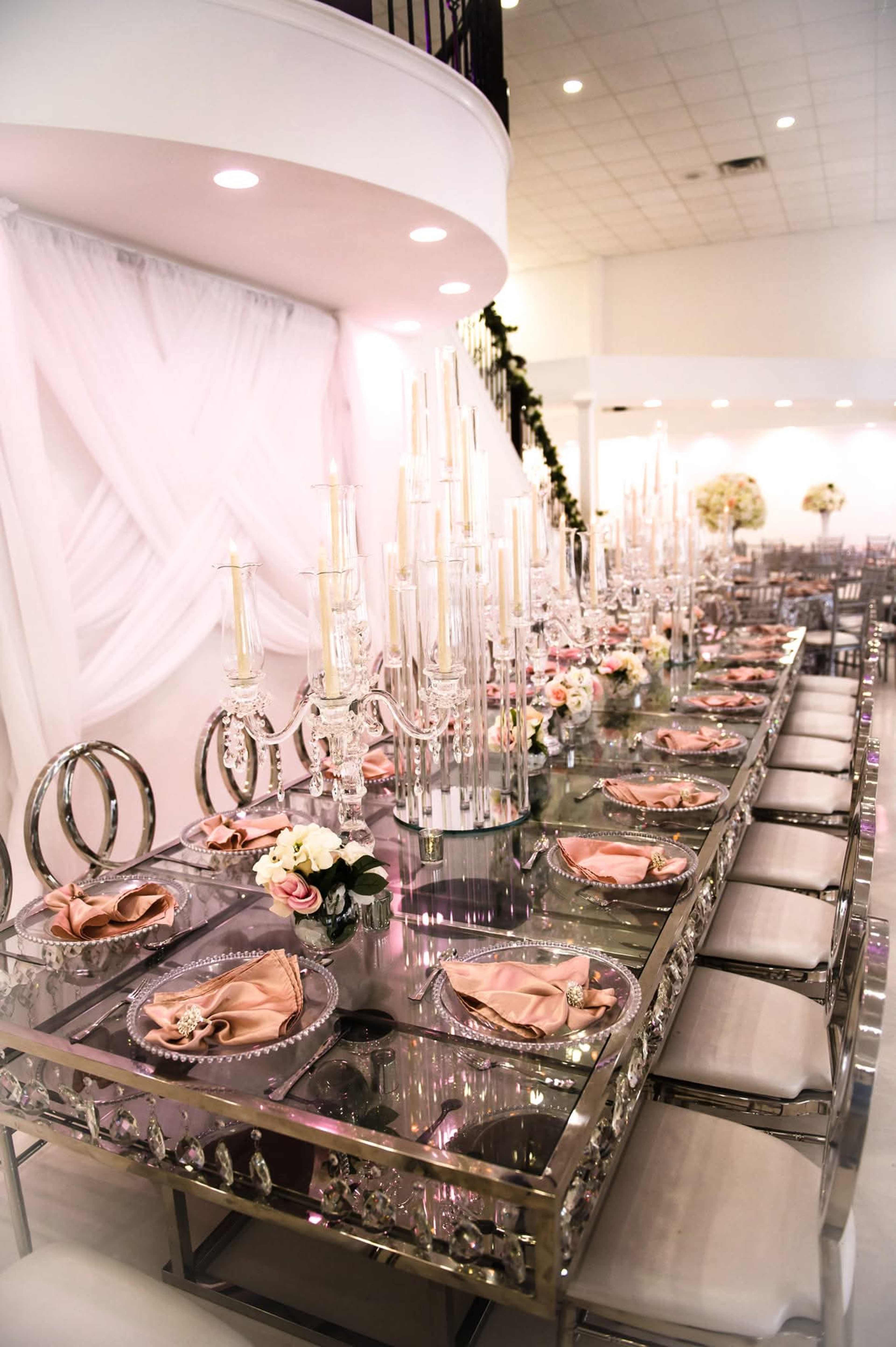 A long, elegant dining table is set with crystal candlesticks, pink napkins, and floral centerpieces, surrounded by decorative chairs in a well-lit banquet hall.