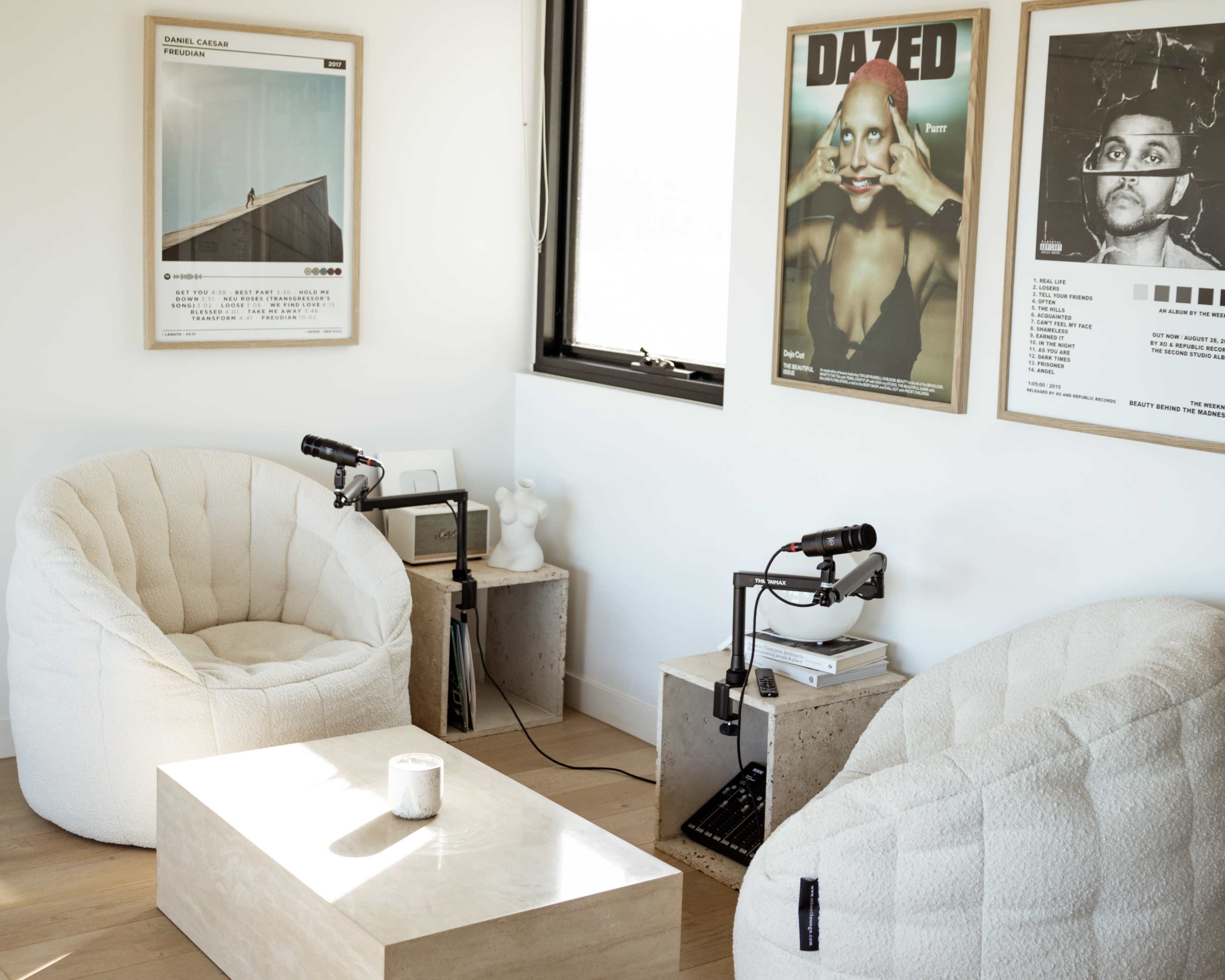 The room features two beige beanbag chairs opposite a light-colored coffee table, with art posters on the walls and microphones on side tables.