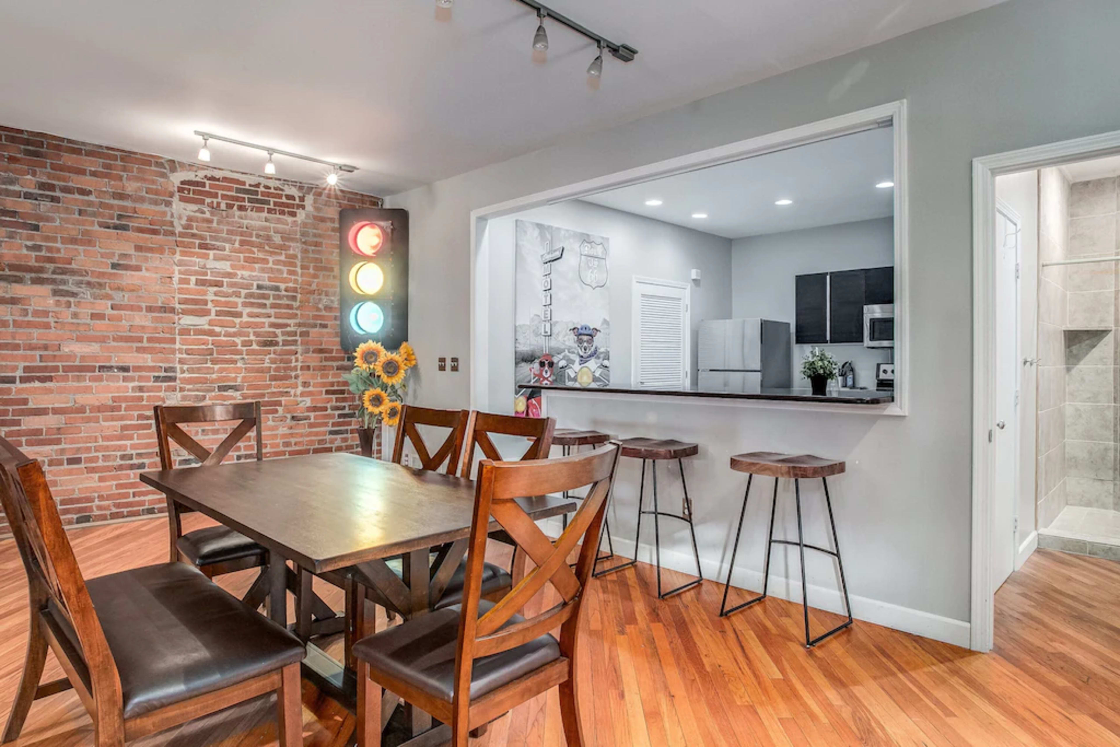 A dining area features a wooden table surrounded by chairs, with a brick wall and a colorful traffic light display in the background.