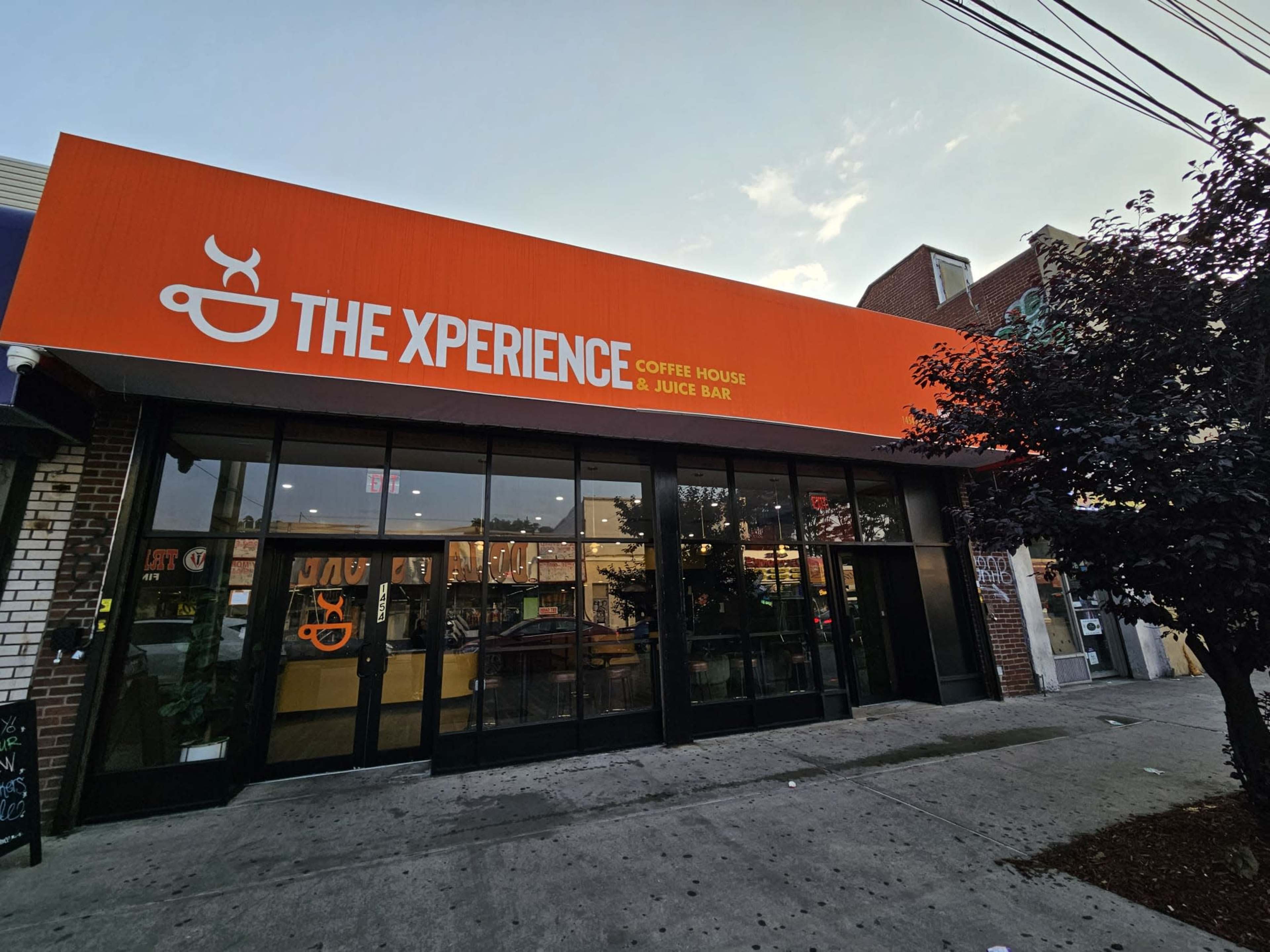 The front facade of "The Xperience" coffee house and juice bar, featuring a bright orange awning and large glass windows.