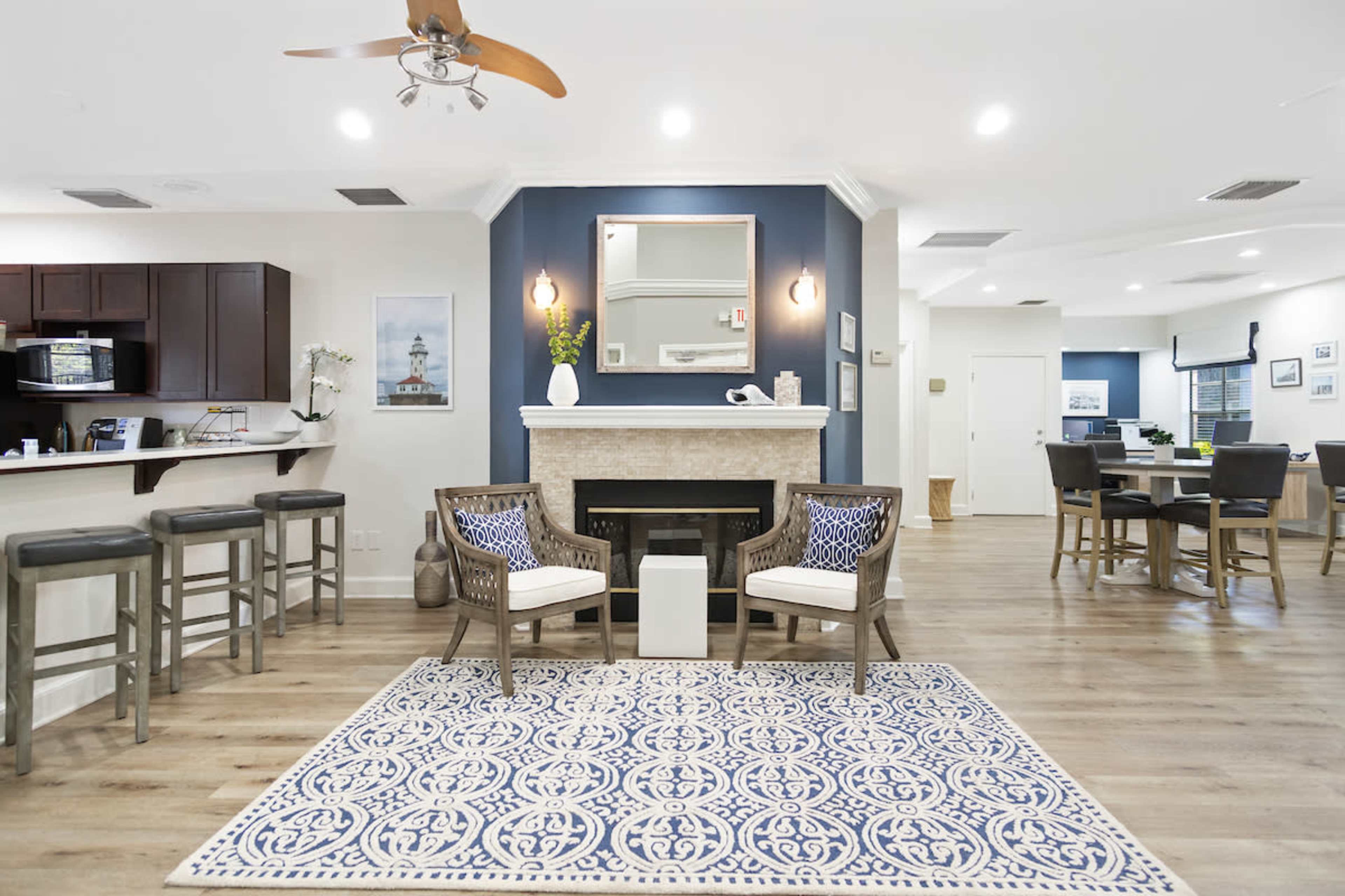 A modern living area with a fireplace, two chairs, and a patterned rug, connected to a dining space and kitchen with dark cabinetry.