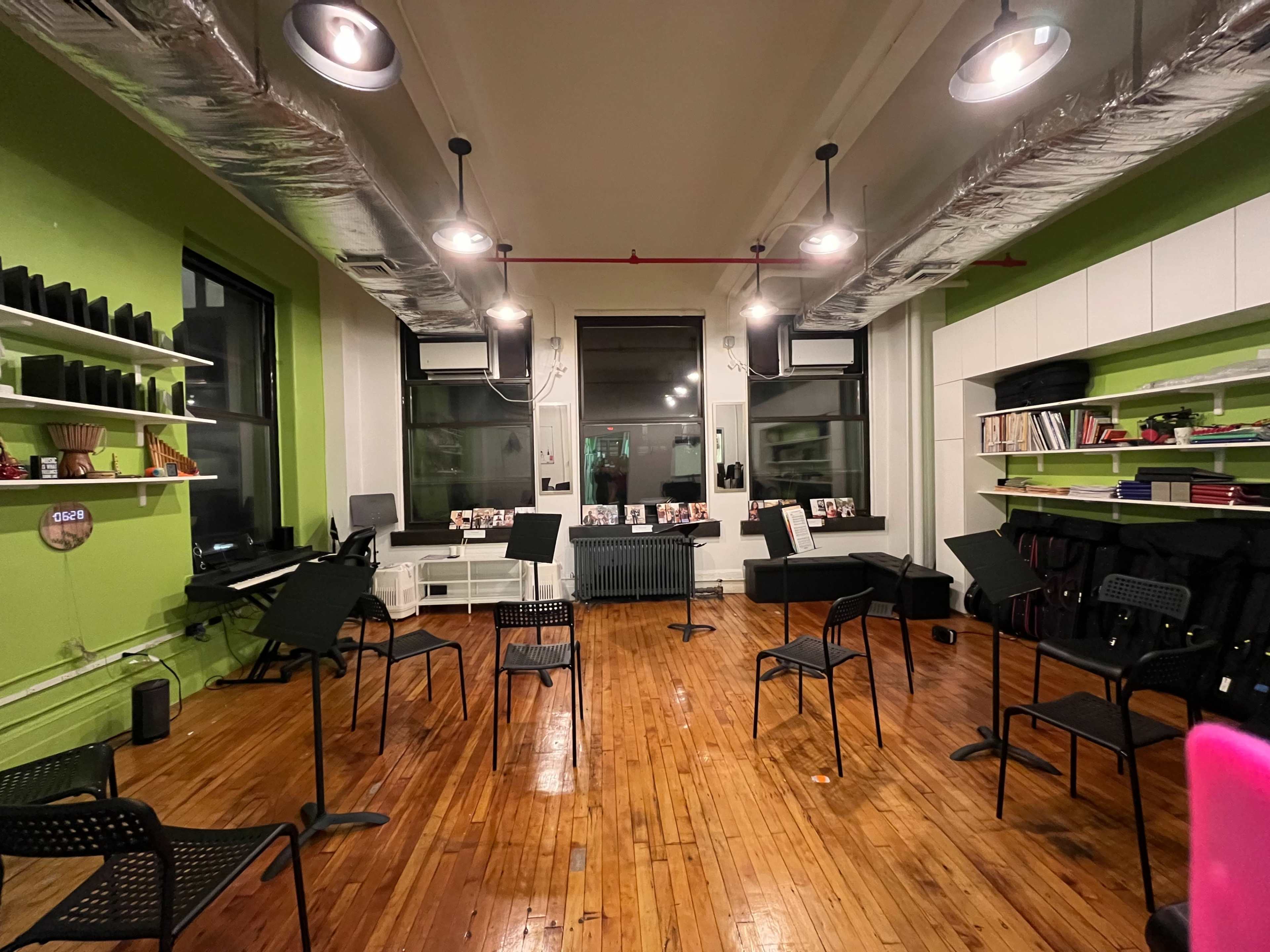 The image shows a rehearsal space with black music stands arranged in a circular formation on a hardwood floor, surrounded by green walls and equipped with shelves containing music books and instruments.