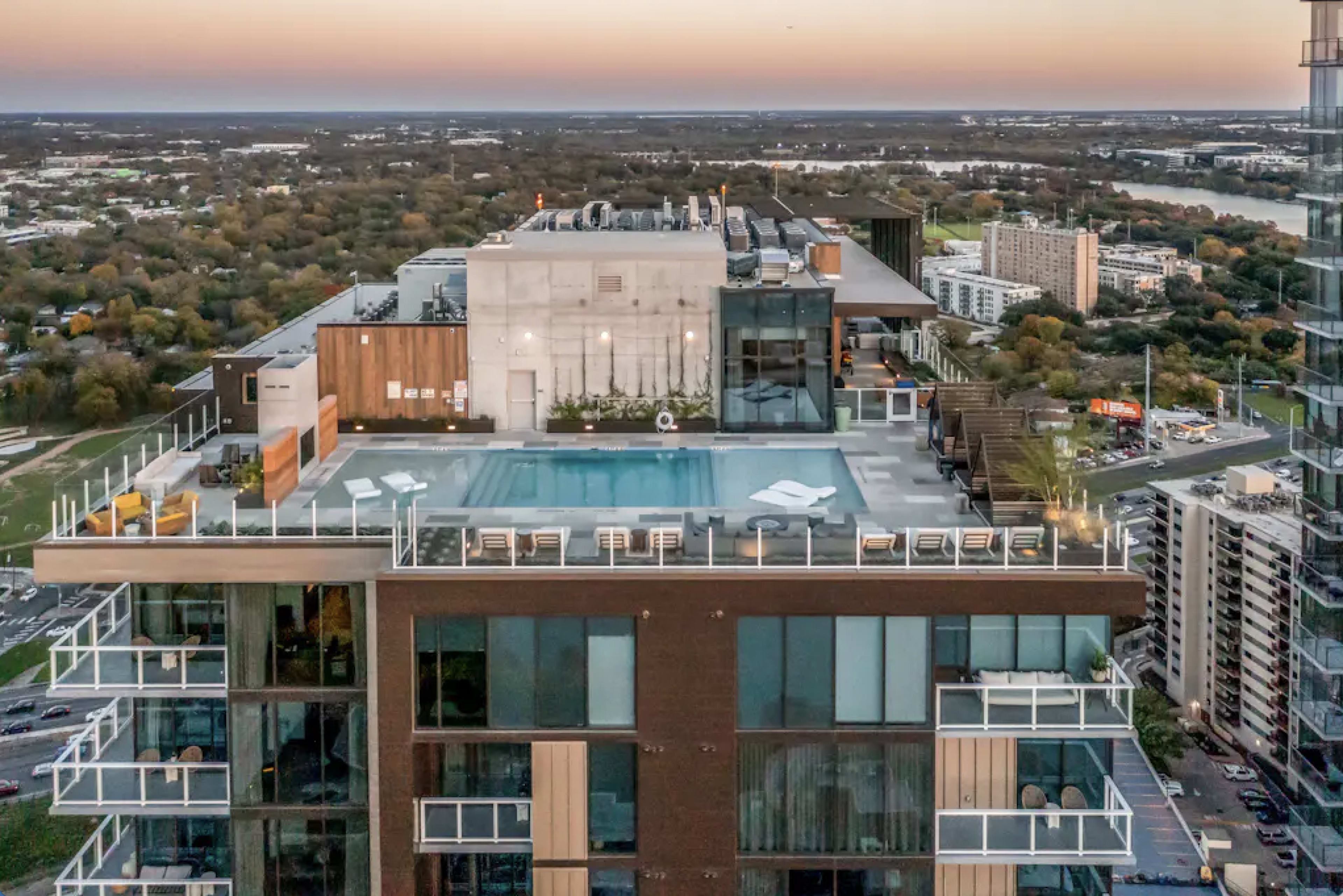 An aerial view shows a modern high-rise building with a rooftop pool and lounge area at sunset.