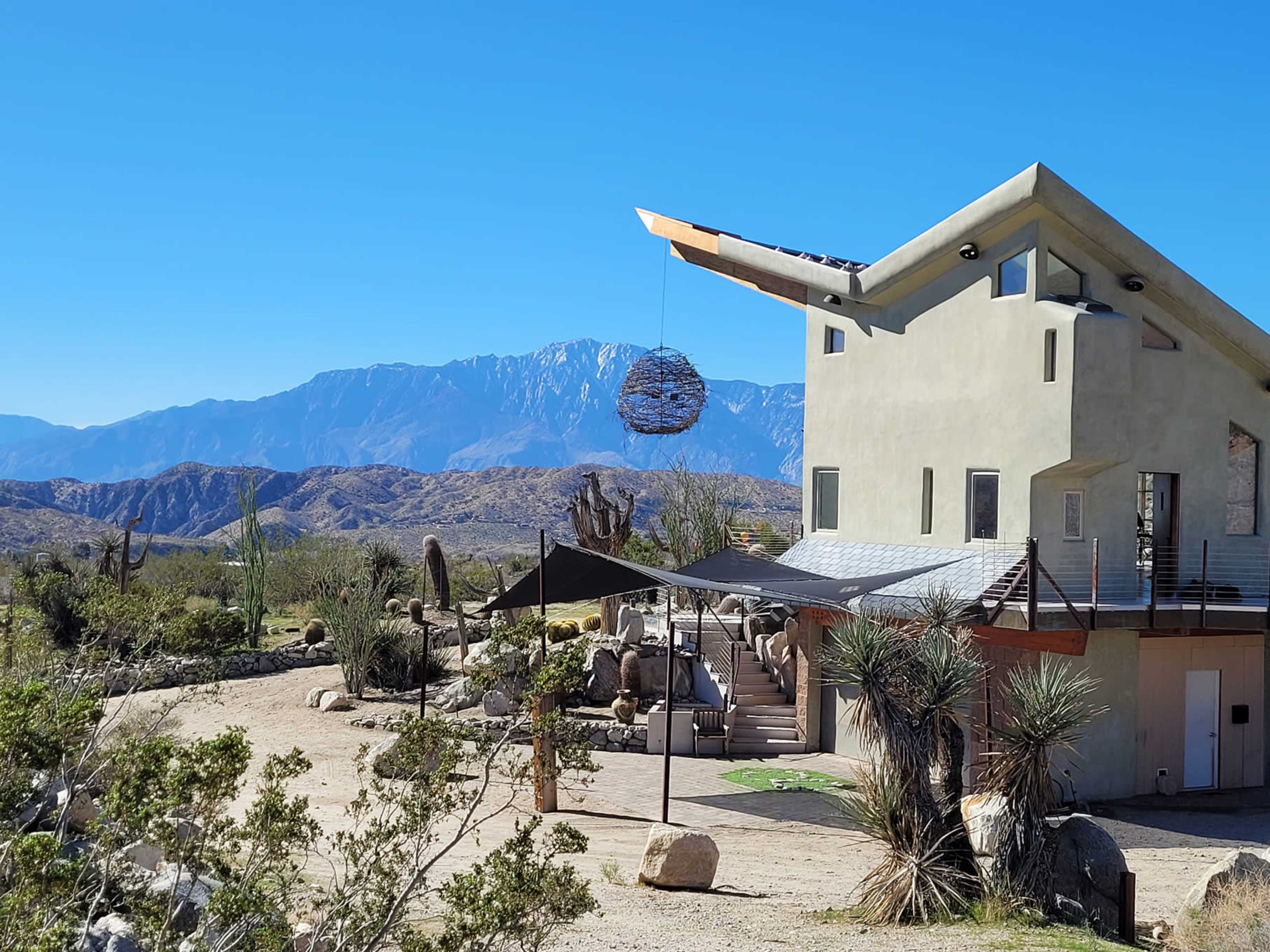 A modern house with an angular design is situated in a desert landscape, with mountains visible in the background and a sculpture hanging above an entrance.