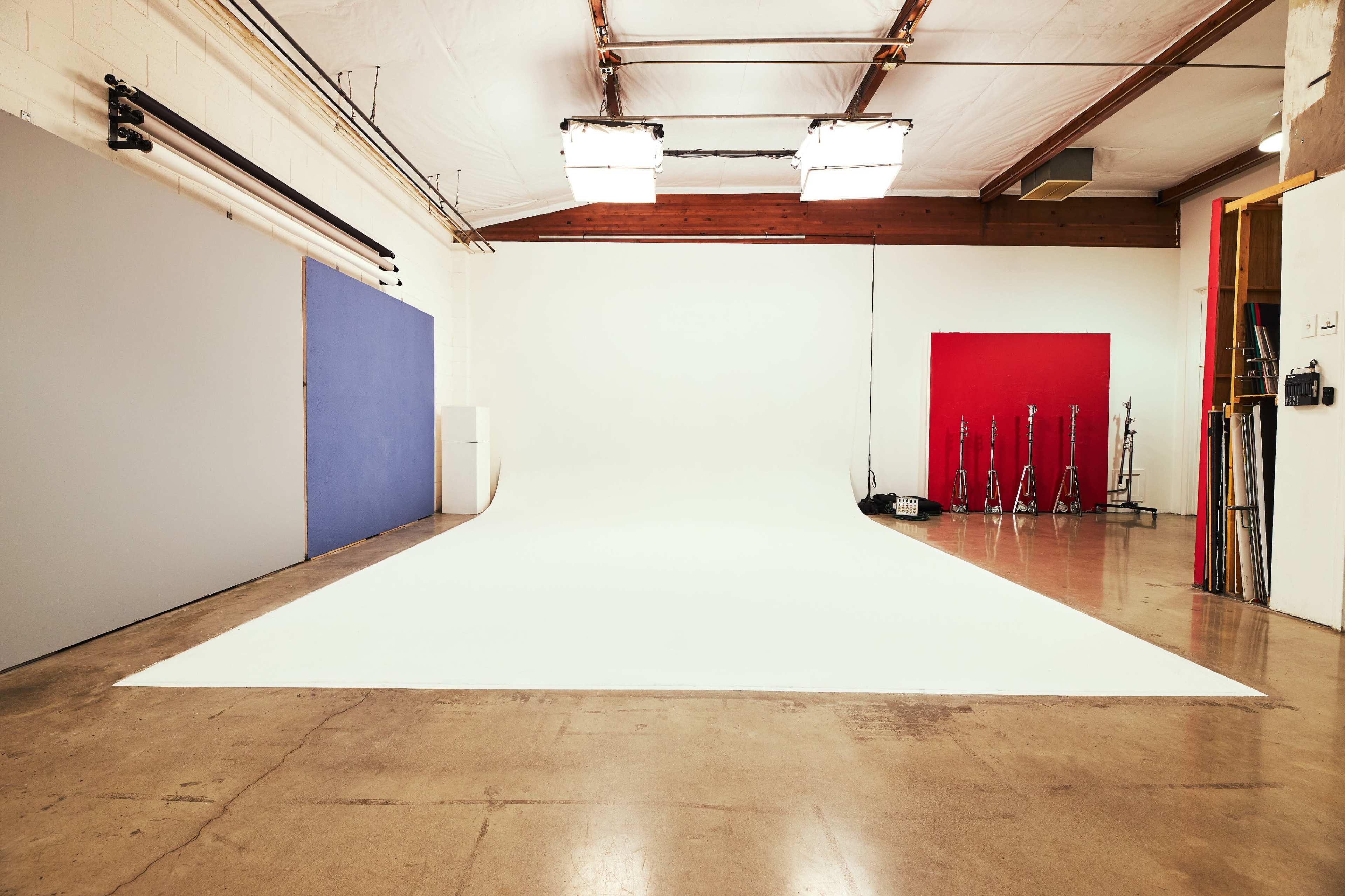 A spacious photography studio with a white seamless backdrop, colored backdrops on the sides, and lighting equipment in the background.