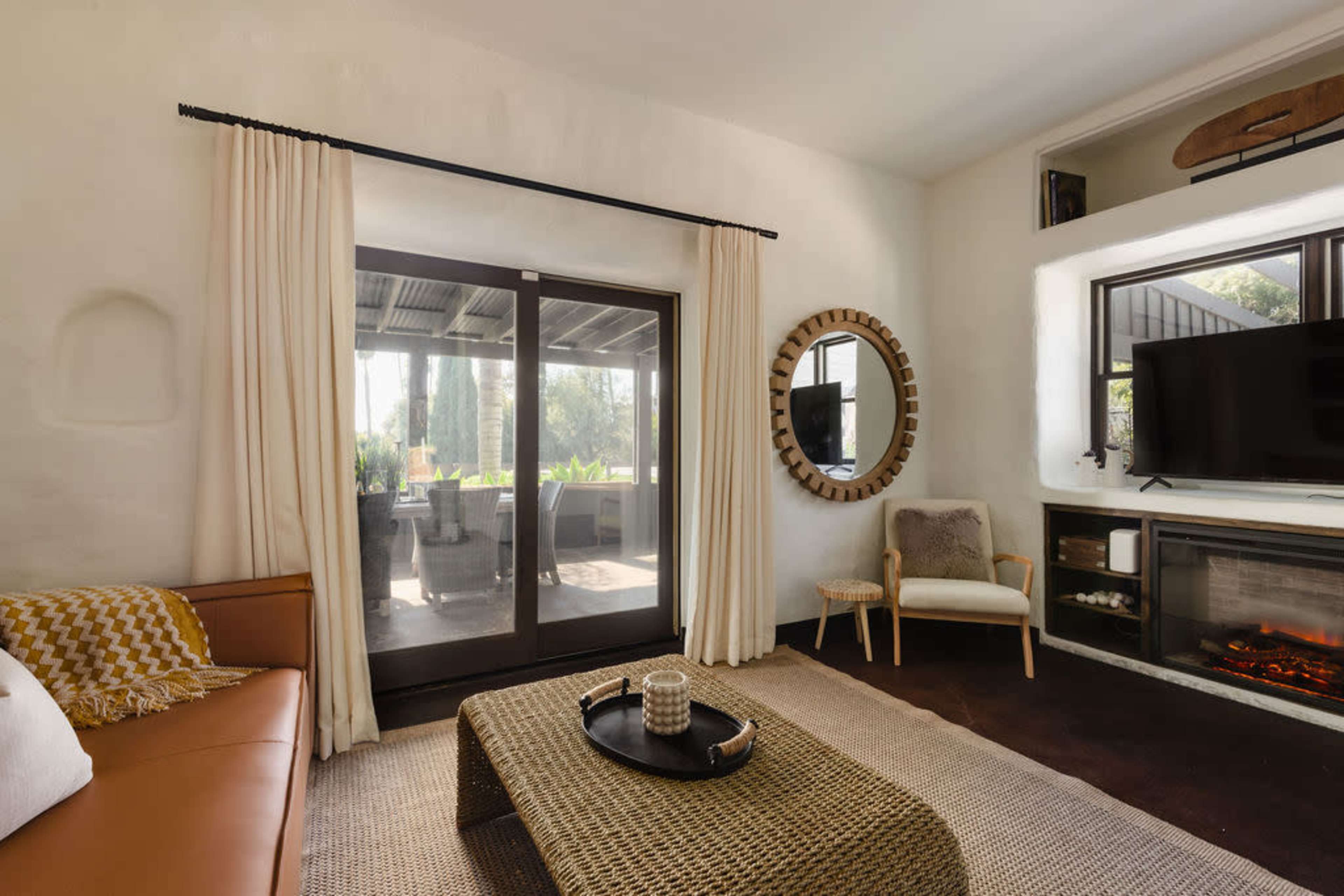 A cozy living room with a leather sofa, a round mirror, a television mounted on the wall, and large glass doors leading to an outdoor patio.