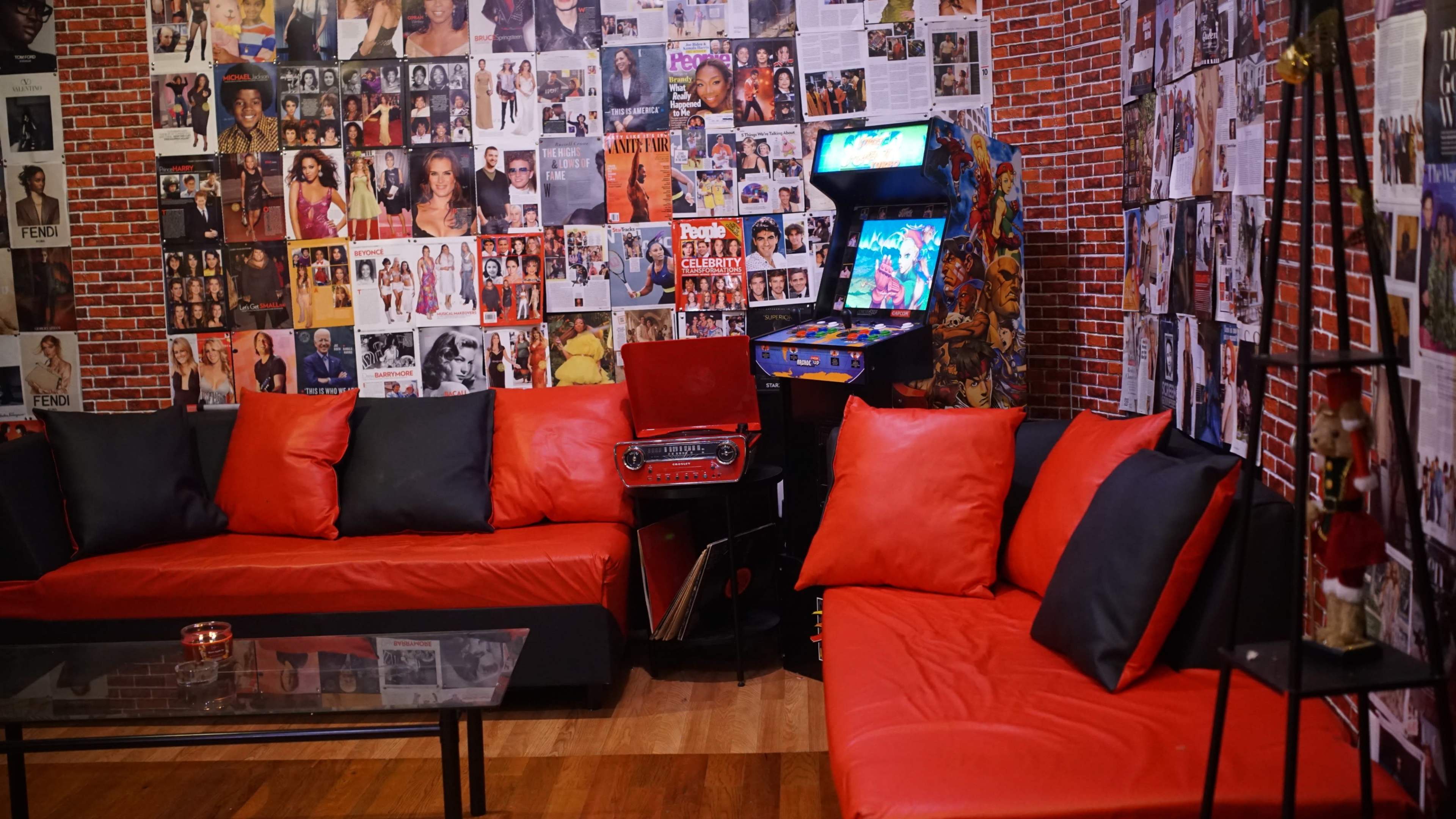 The scene features a retro-themed lounge area with red and black sofas, a glass coffee table, and walls adorned with various photographs, alongside a vintage arcade machine in the corner.