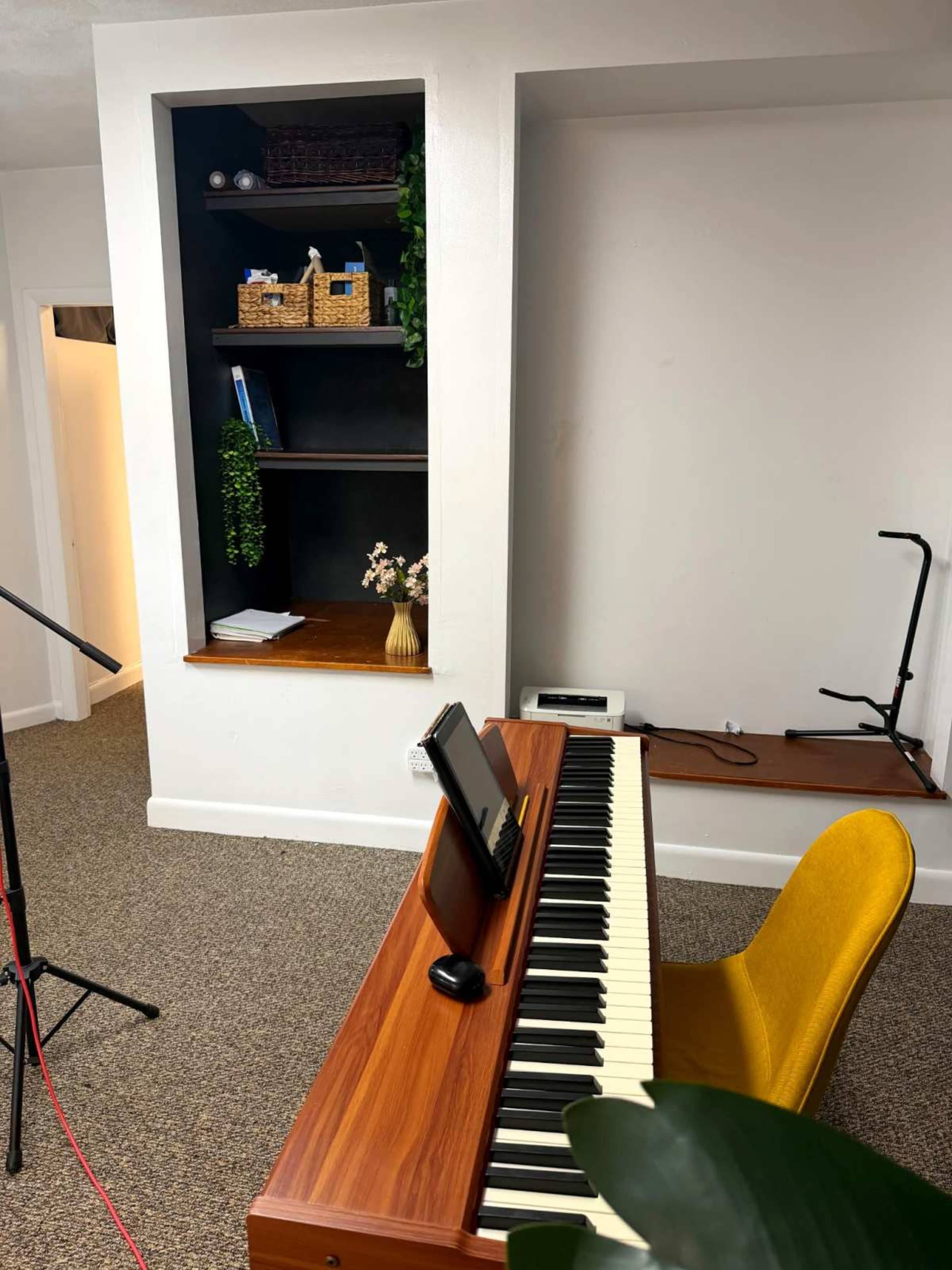 A digital piano is positioned in front of a small bookshelf built into the wall, which holds decorative items and plants.