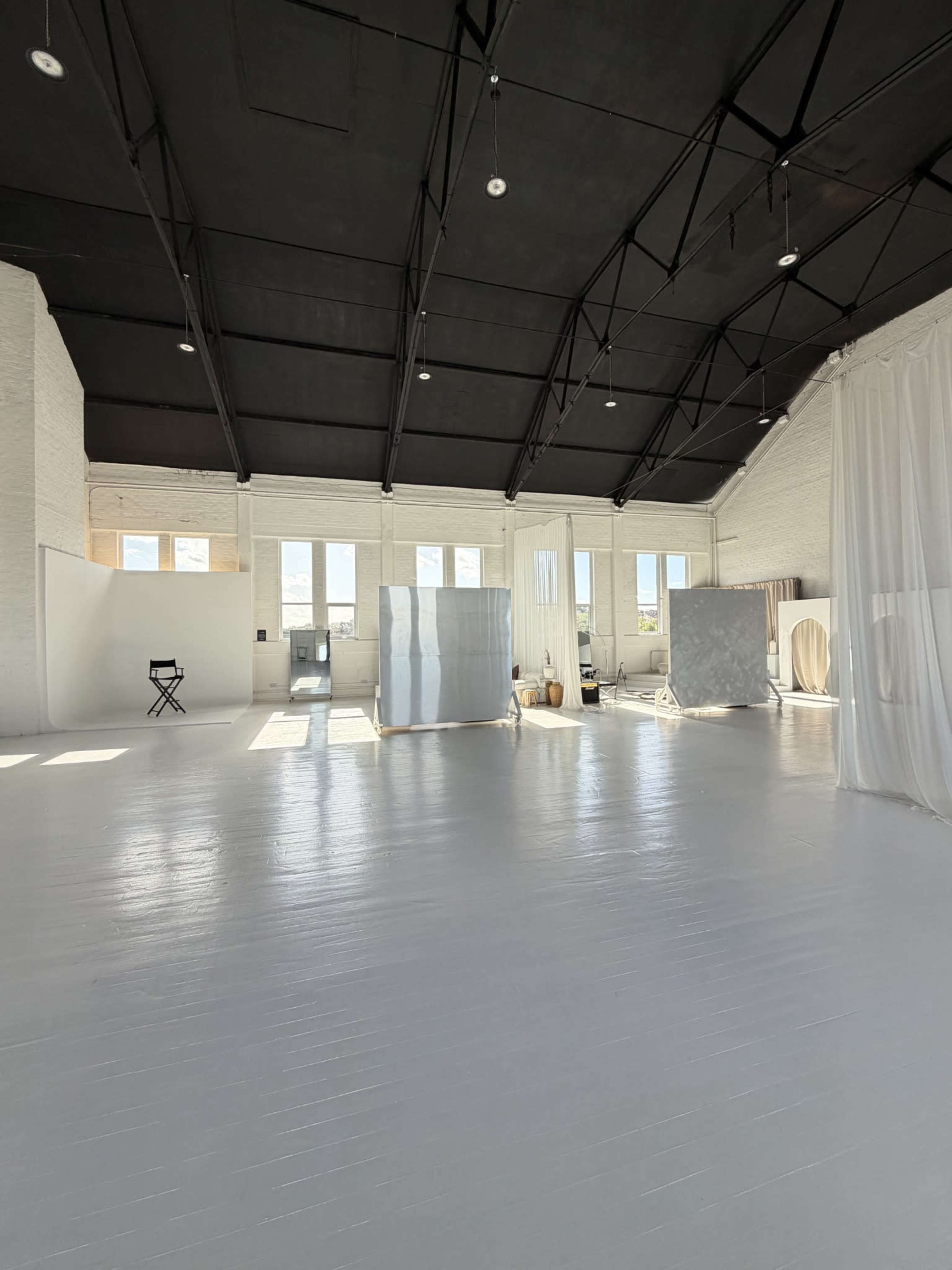 The image shows a spacious, well-lit studio with large windows, white walls, and several fabric dividers creating distinct areas.