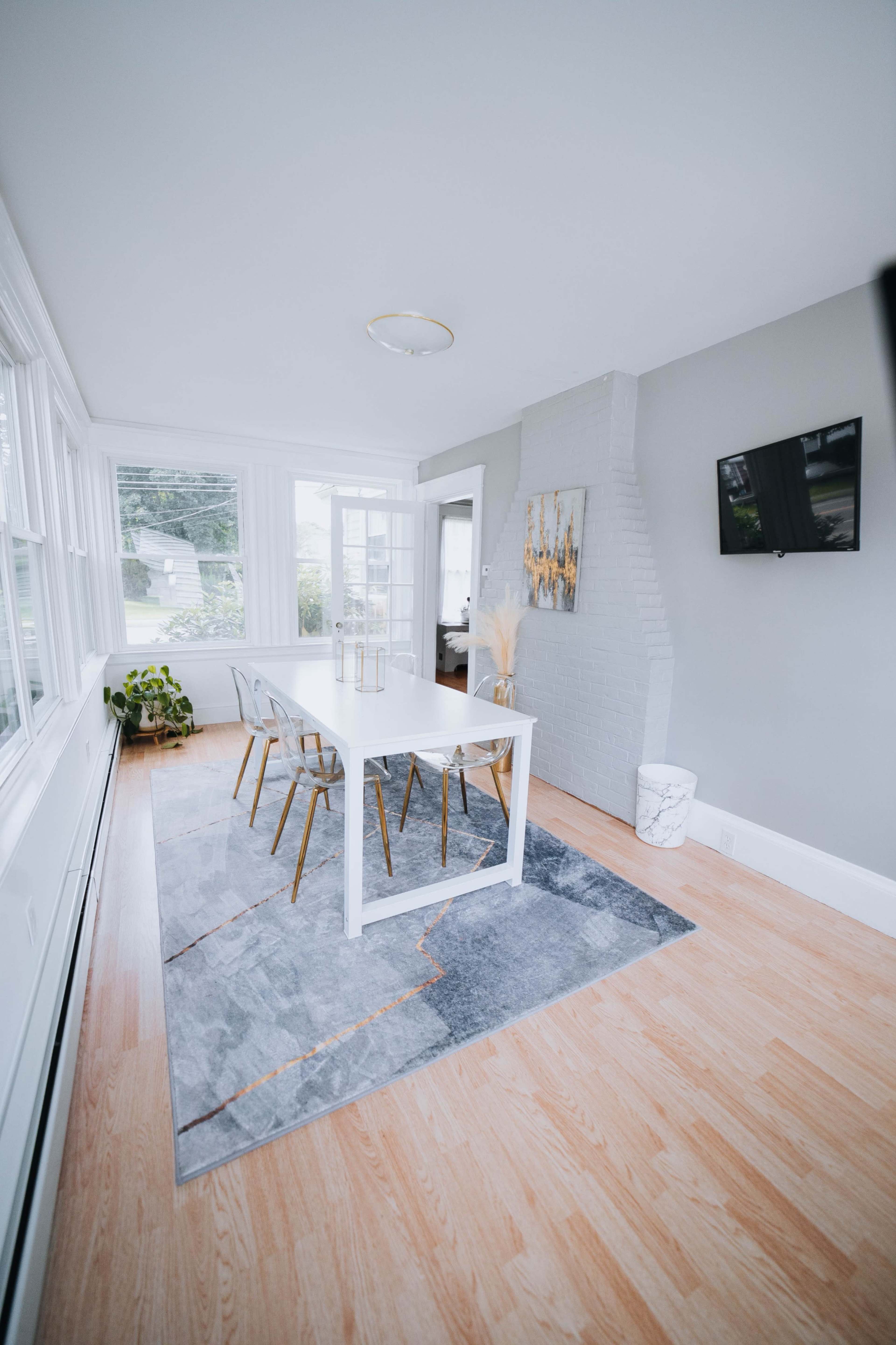 A bright dining area featuring a white table surrounded by gold-accented chairs on a gray and orange rug, with large windows allowing natural light.
