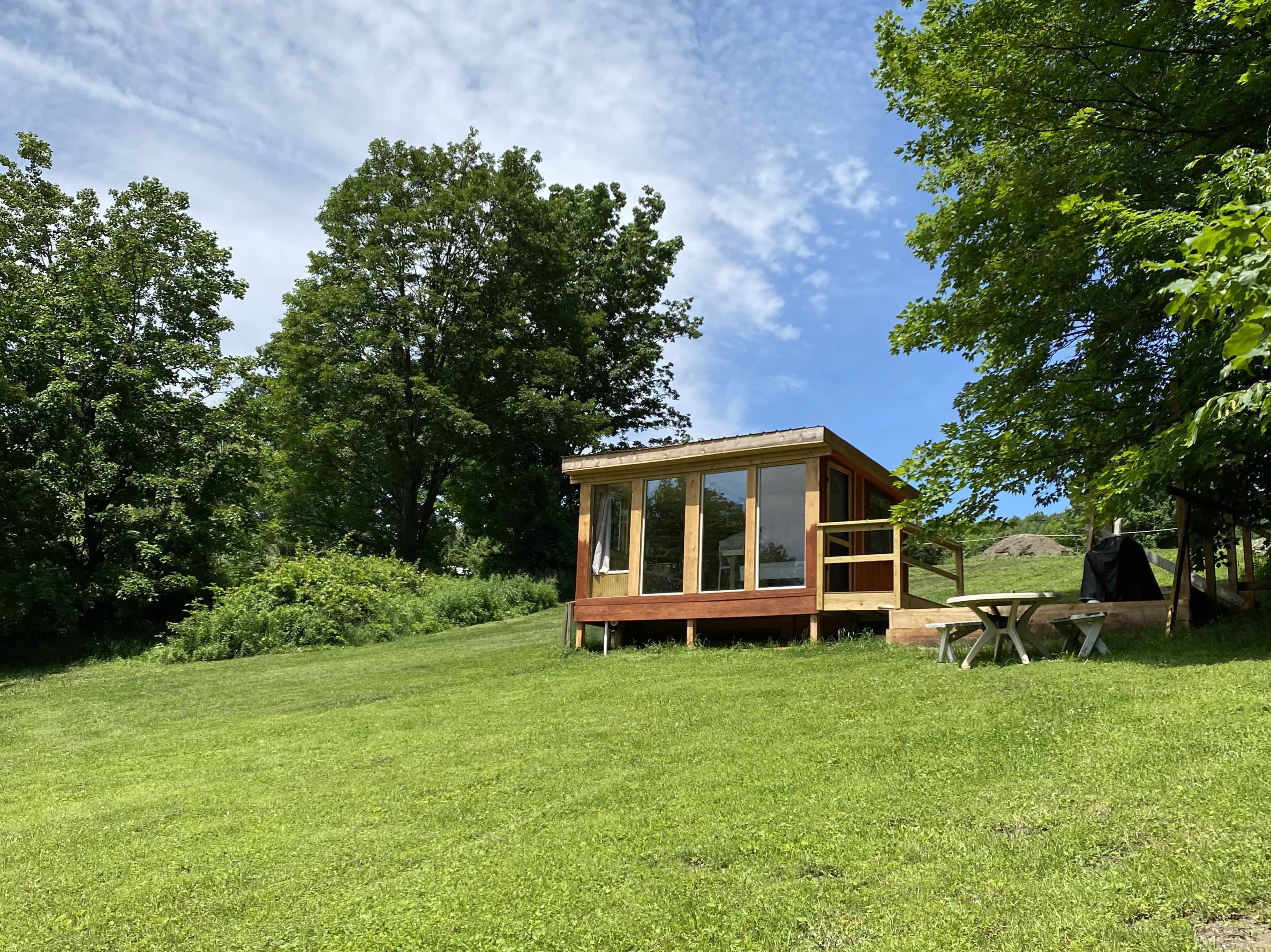 A small cabin with large windows sits on a grassy slope surrounded by trees.