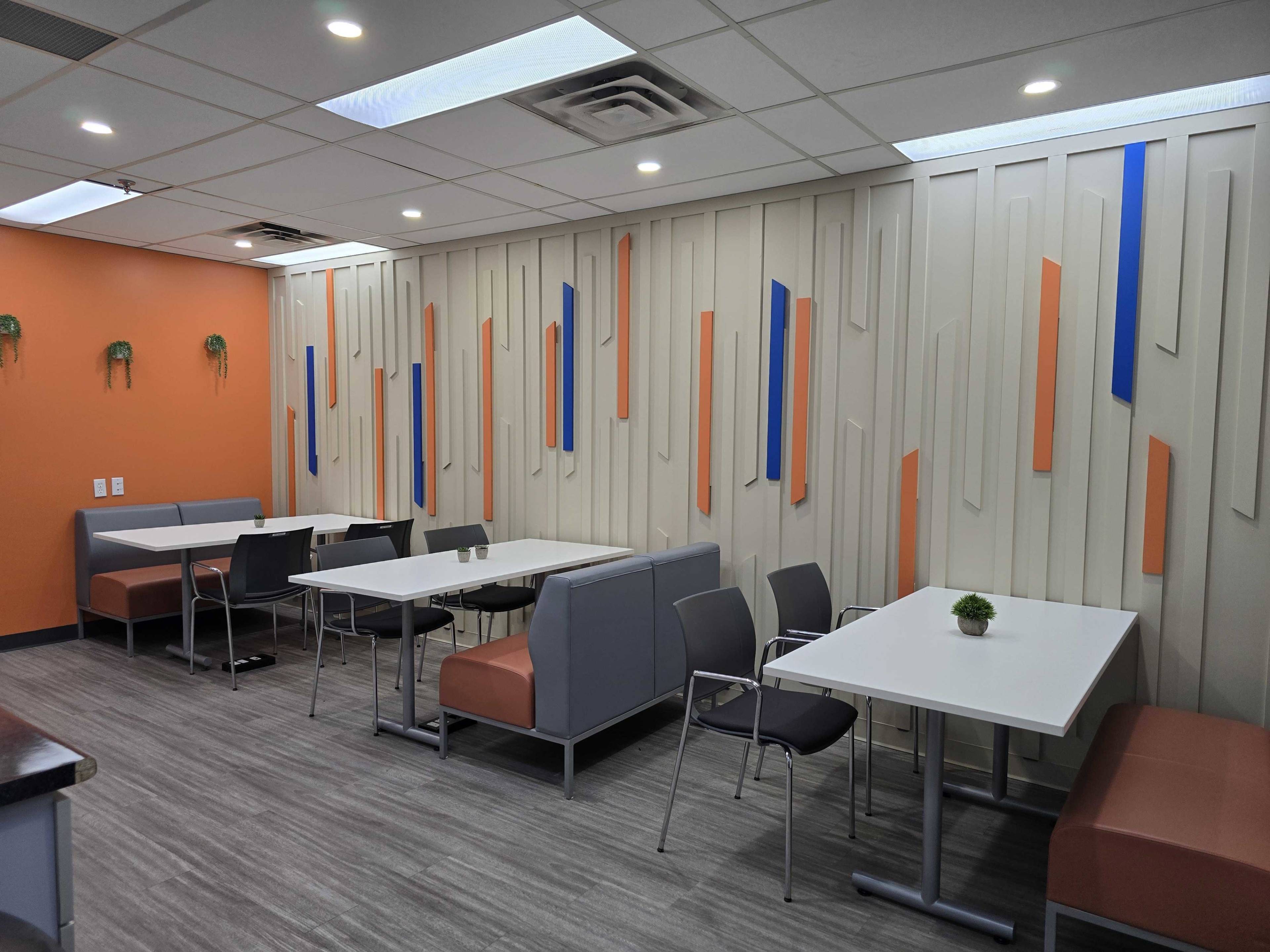 The image shows a modern dining area with white tables, gray and orange seating, and an accent wall featuring abstract architectural elements.