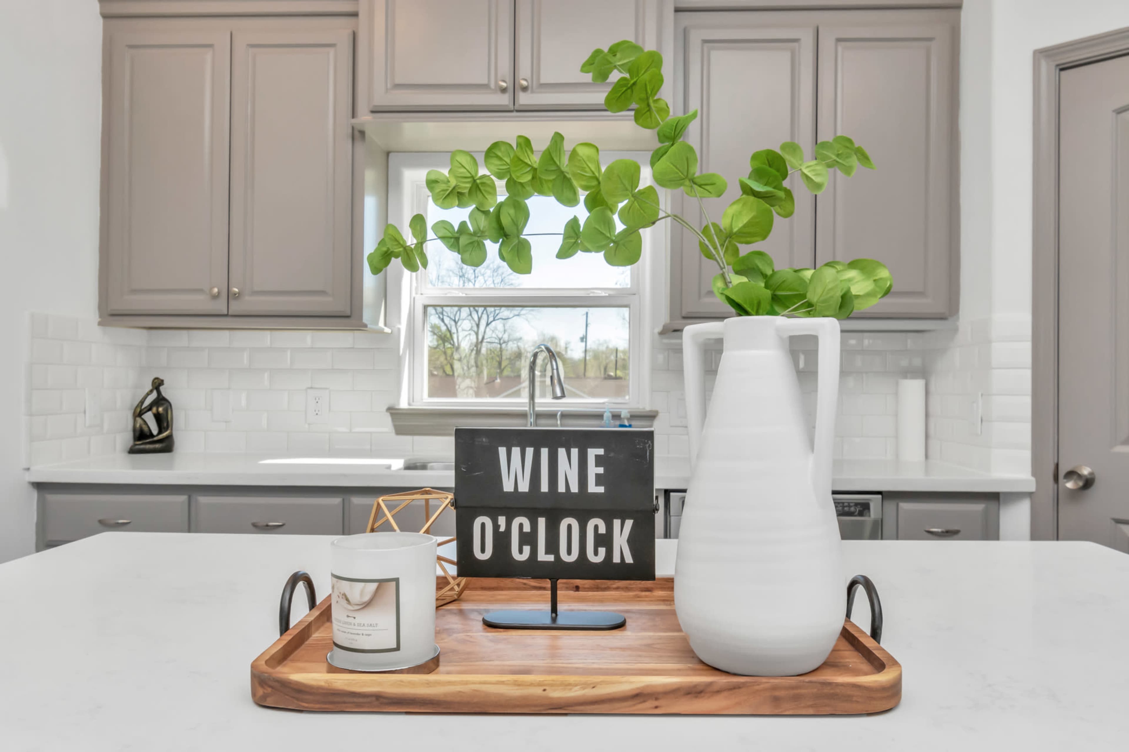 A wooden tray on a kitchen countertop displays a candle, a decorative object, and a vase with green foliage, alongside a sign that reads "WINE O'CLOCK."
