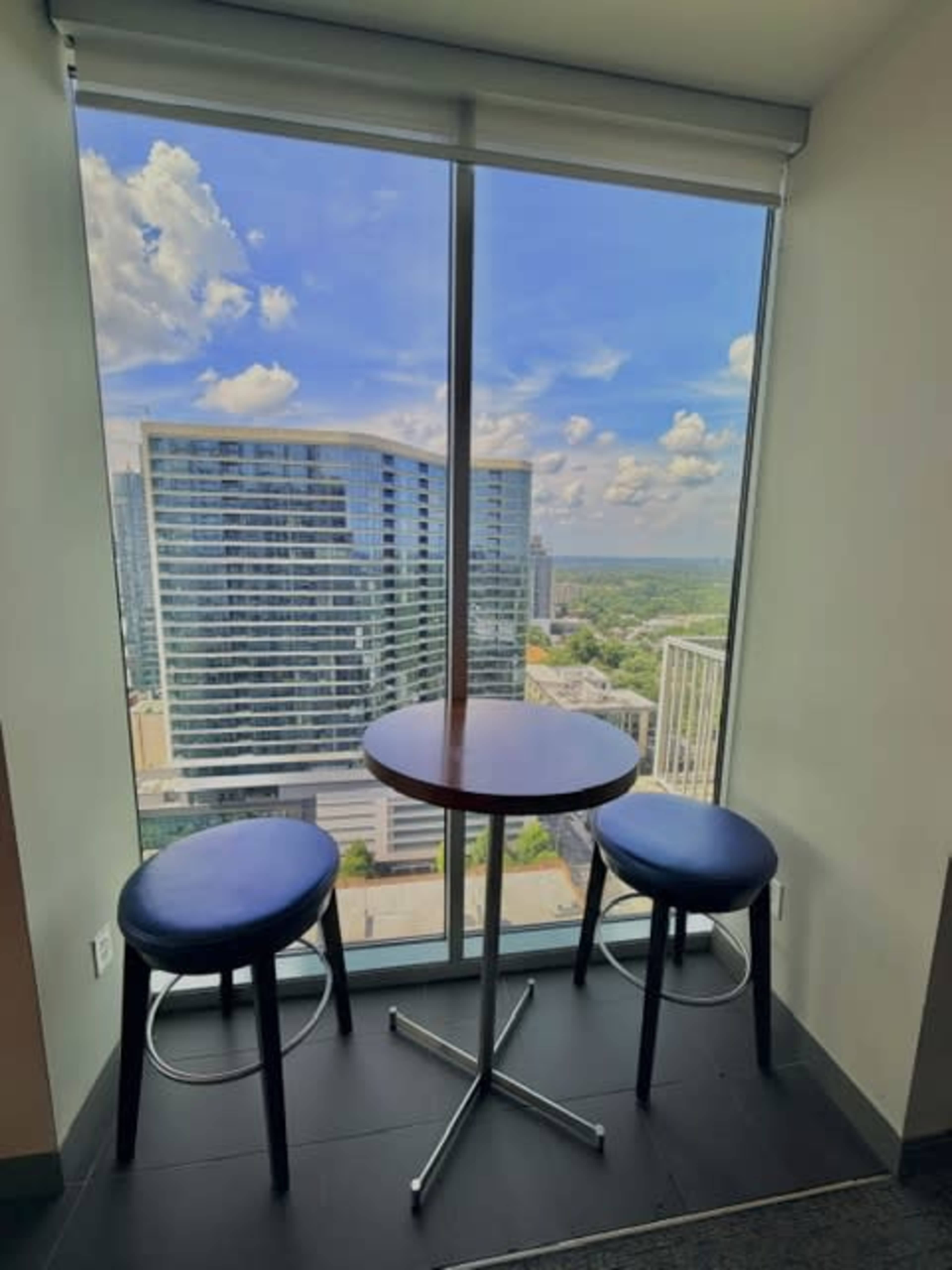 A small round table with two blue stools sits by a large window overlooking a city skyline with tall buildings and a blue sky.