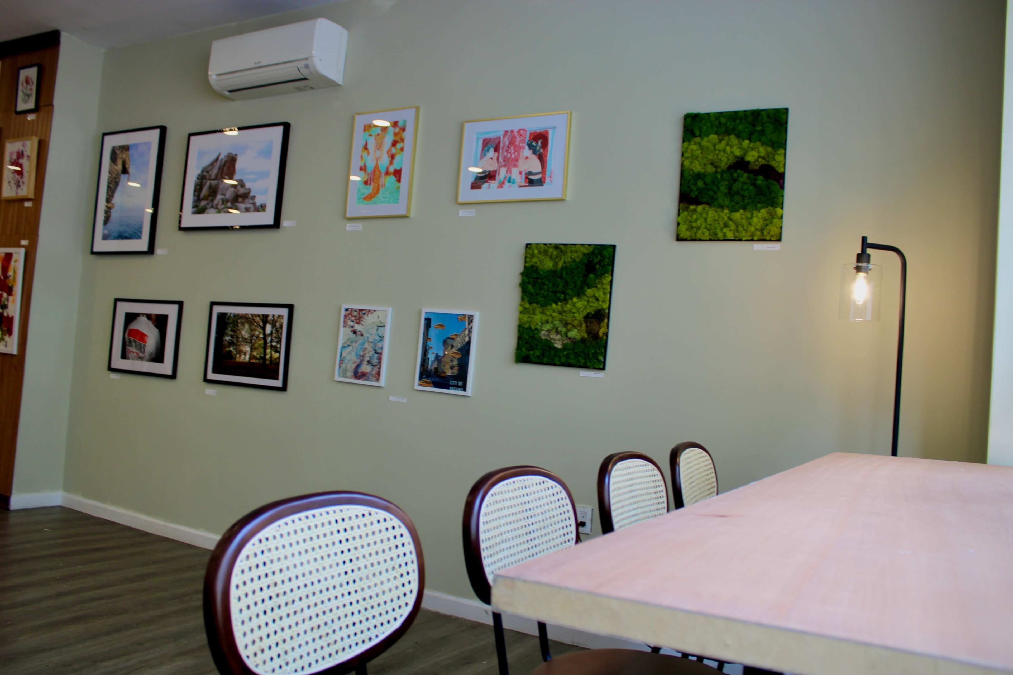 A corner of a gallery or café featuring a wall adorned with framed artworks and a table with several chairs.