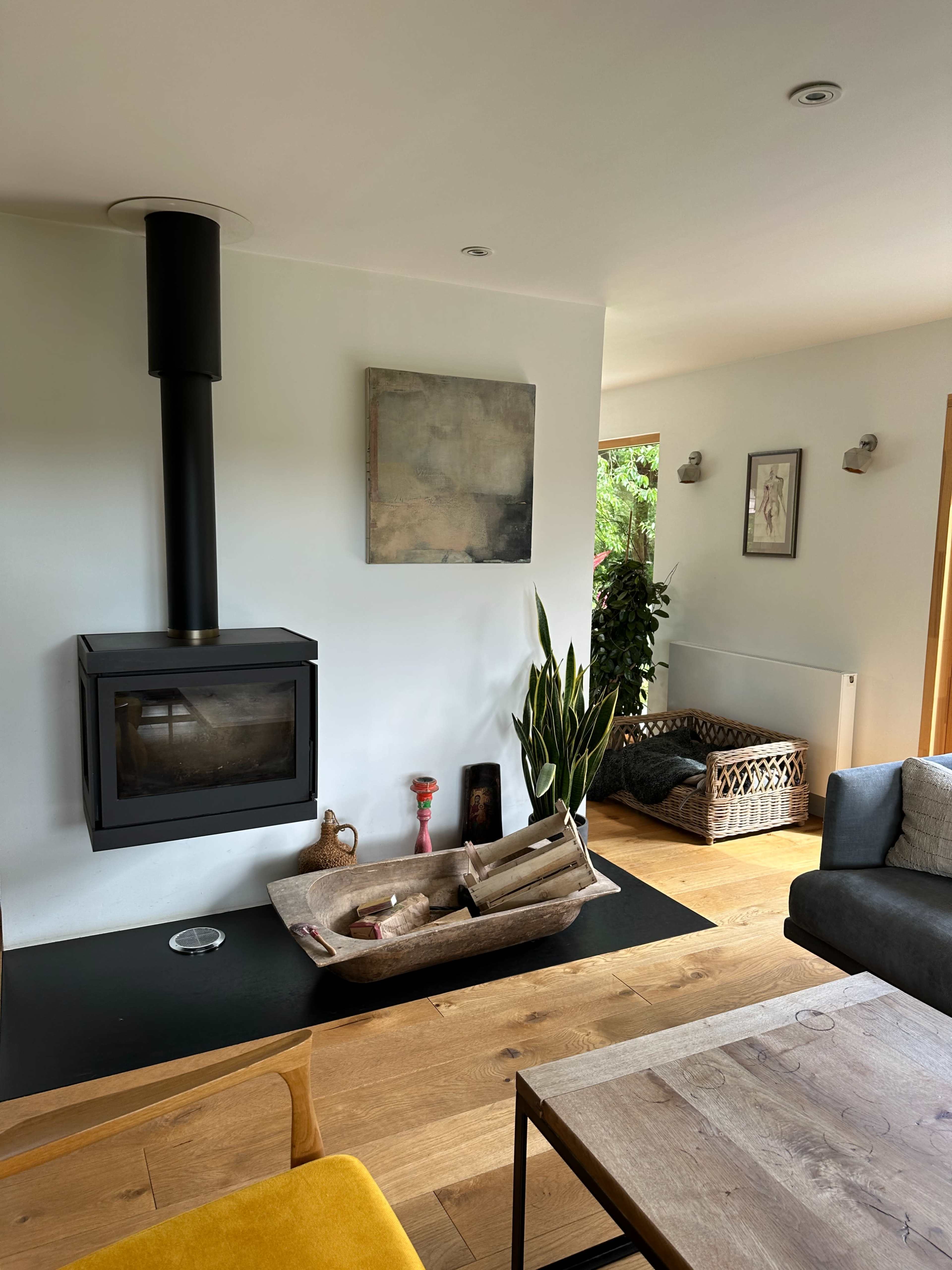 A modern living room with a black wood stove, a large wooden coffee table, and natural light streaming through a nearby window.