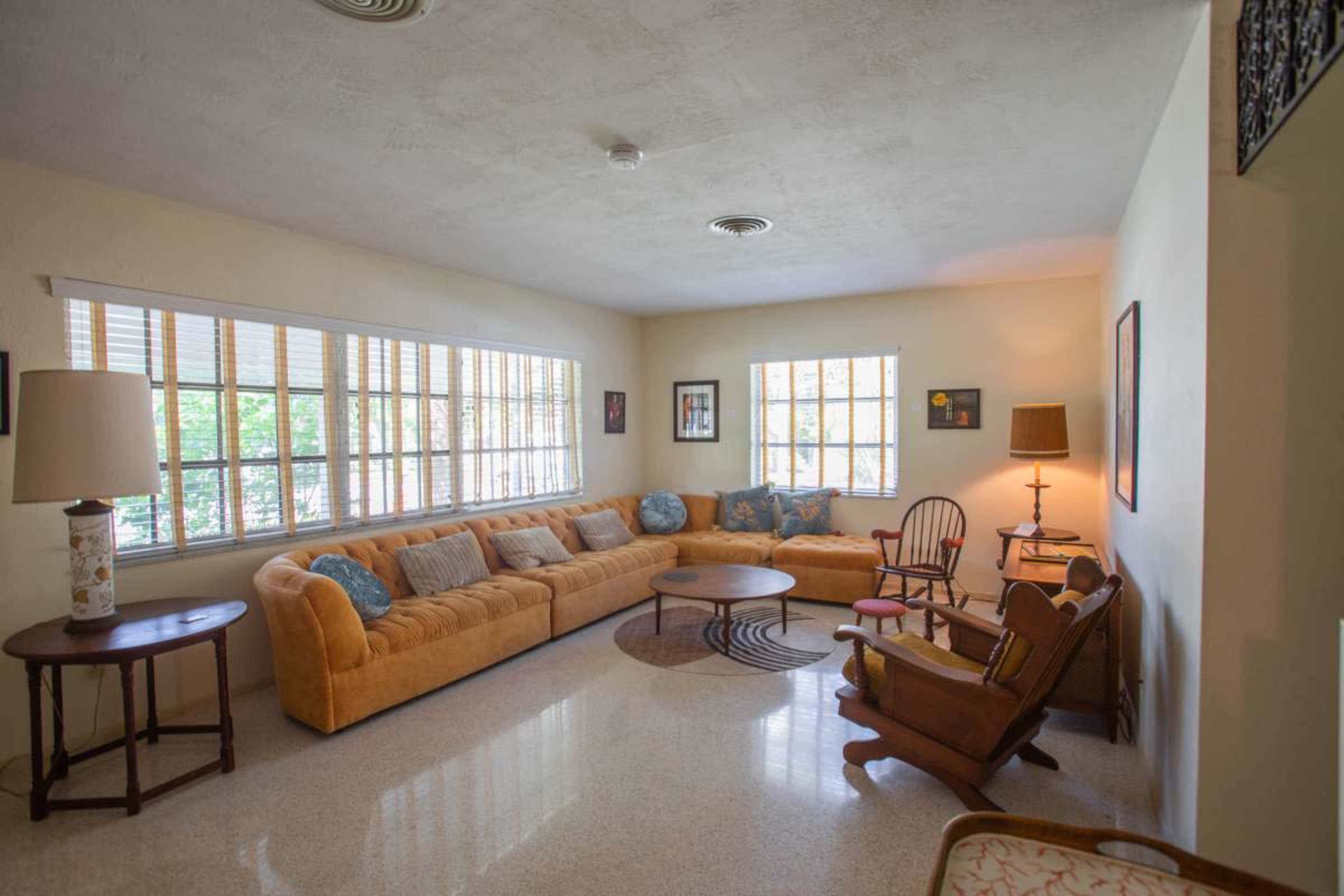 The image shows a spacious living room with a large yellow sectional sofa, a round coffee table, and several chairs arranged around a bright windowed area.
