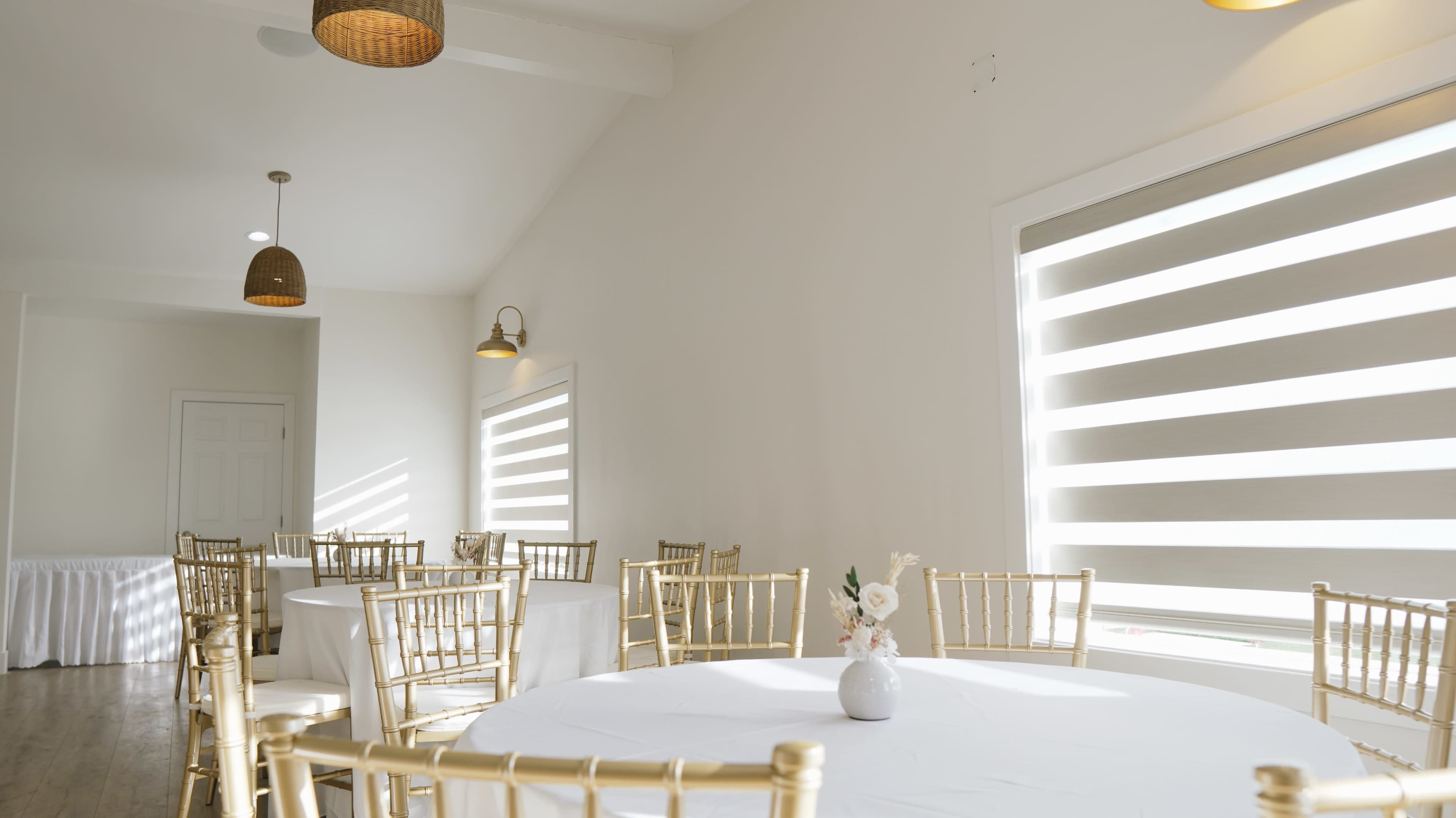 The scene features a bright, modern dining area with gold Chiavari chairs and round tables, illuminated by soft light from large, striped windows.