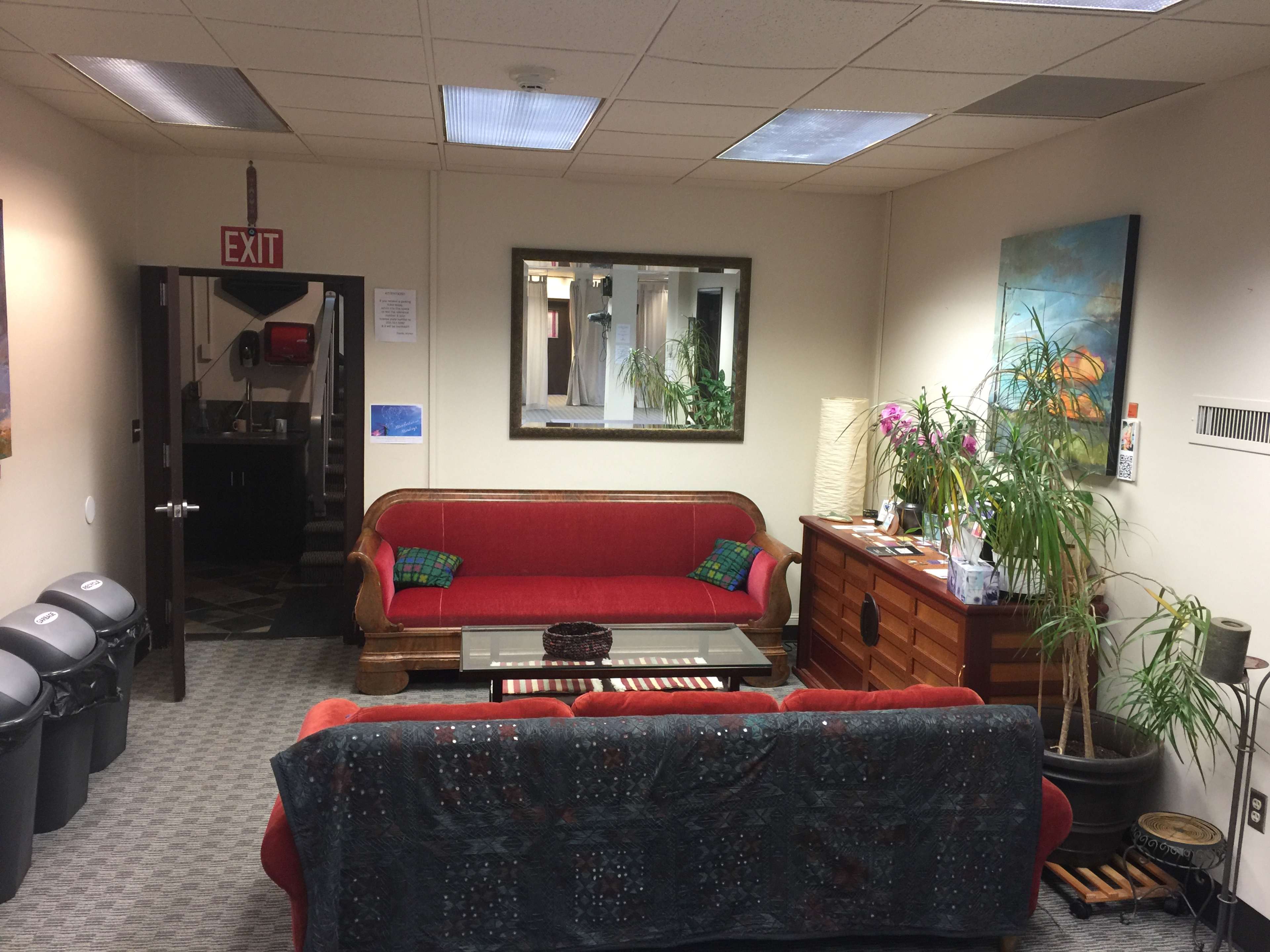 A waiting room with two red couches, a coffee table in the center, a wooden cabinet, and decorative plants, along with an exit sign and trash bins in the background.