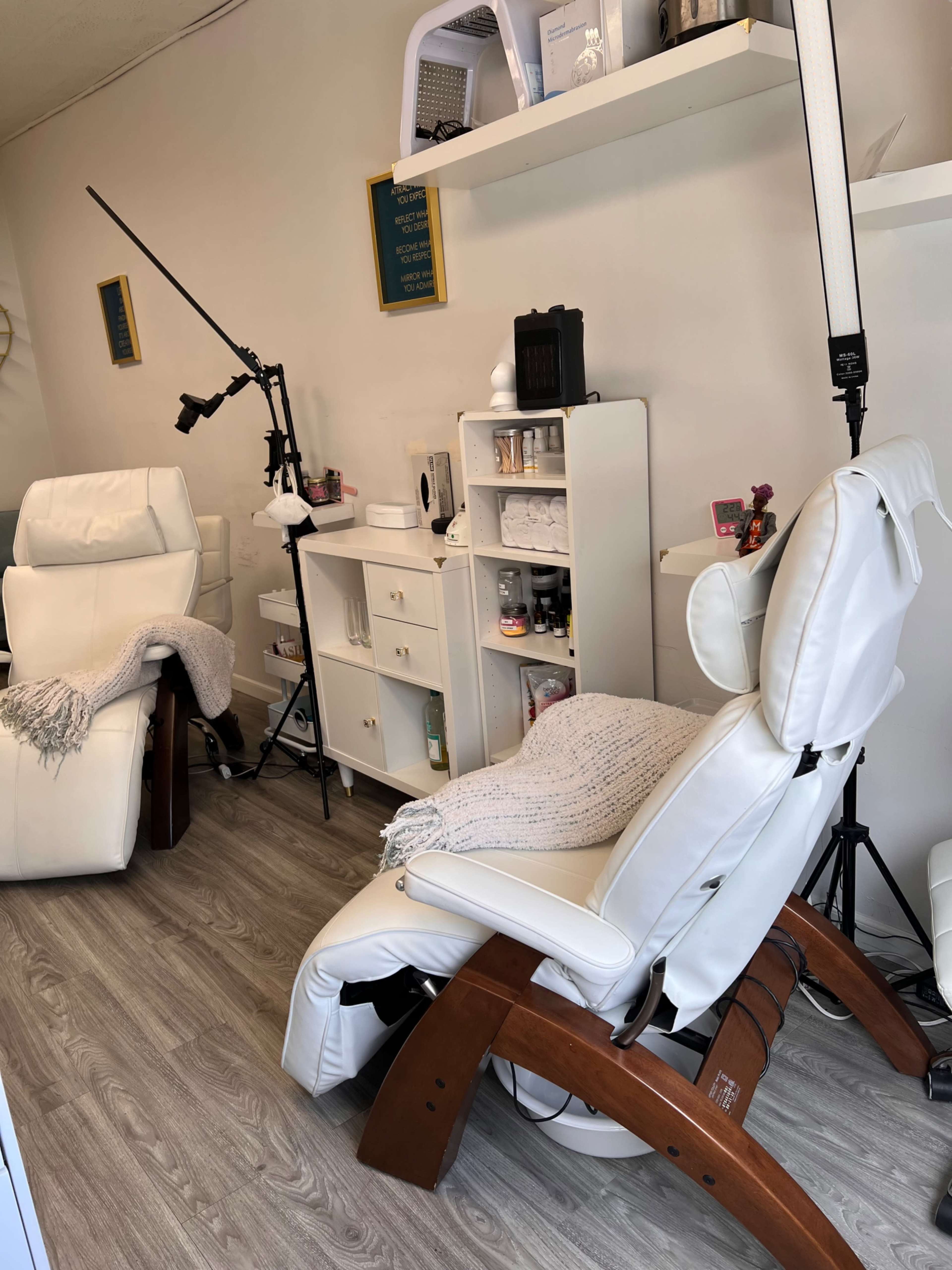The image shows a small spa room featuring a white reclining chair, a nearby storage unit with various beauty products, and a tripod light setup.