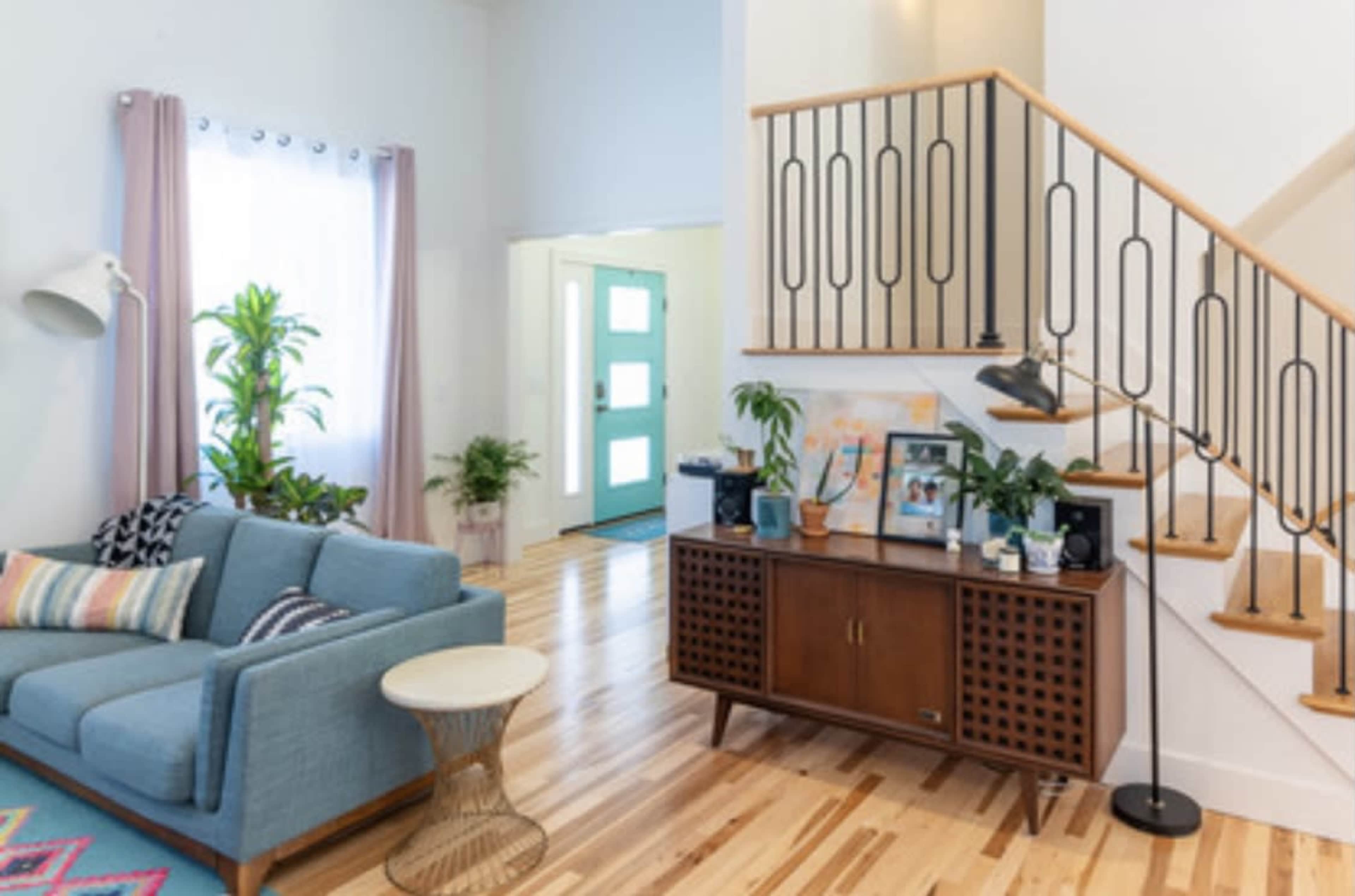 A bright living room with a blue sofa, a wooden side table, potted plants, and a staircase leading to an upper level.