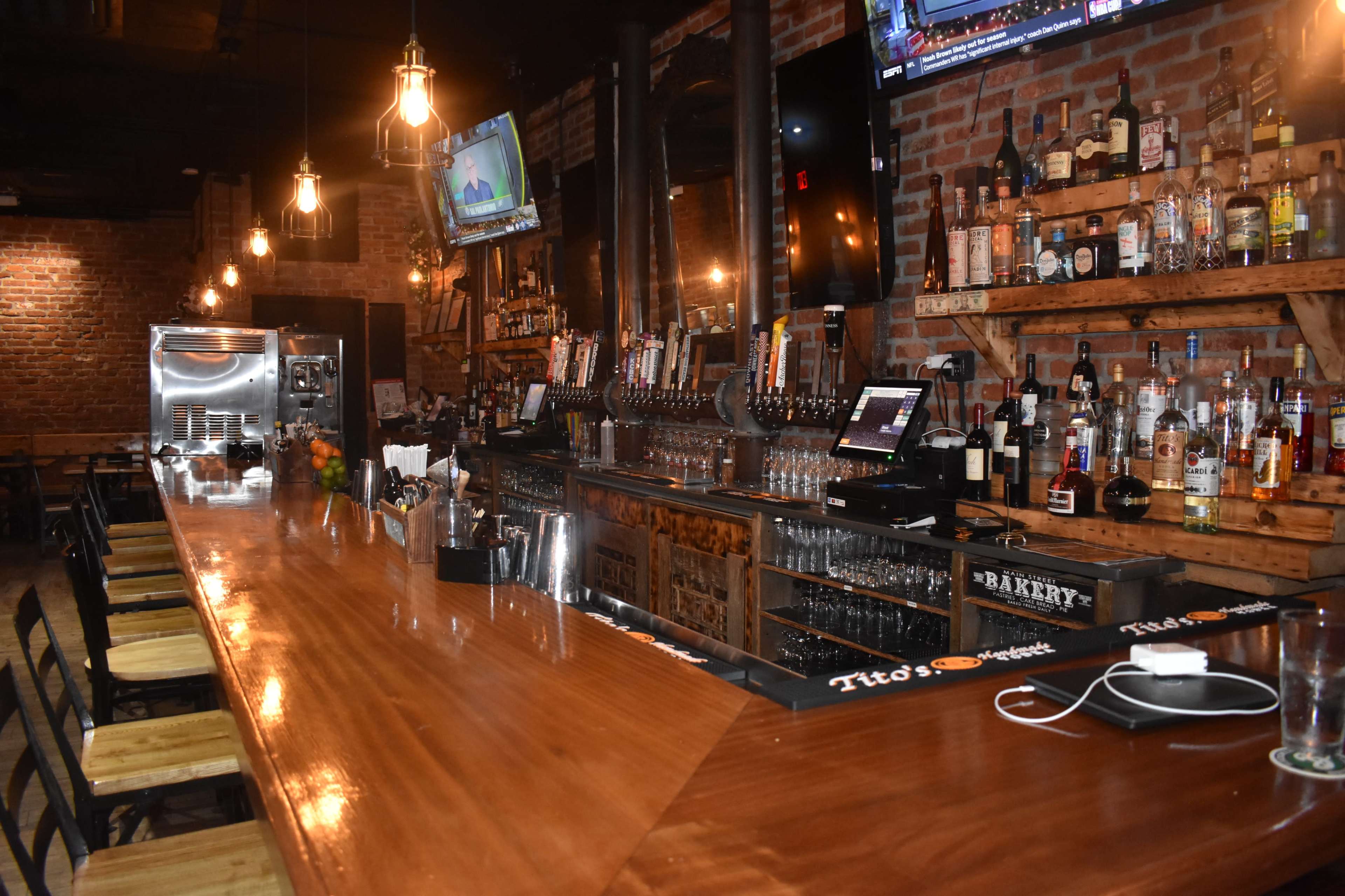 The image shows a rustic bar with a wooden counter, various liquor bottles on shelves, and several television screens mounted on brick walls.