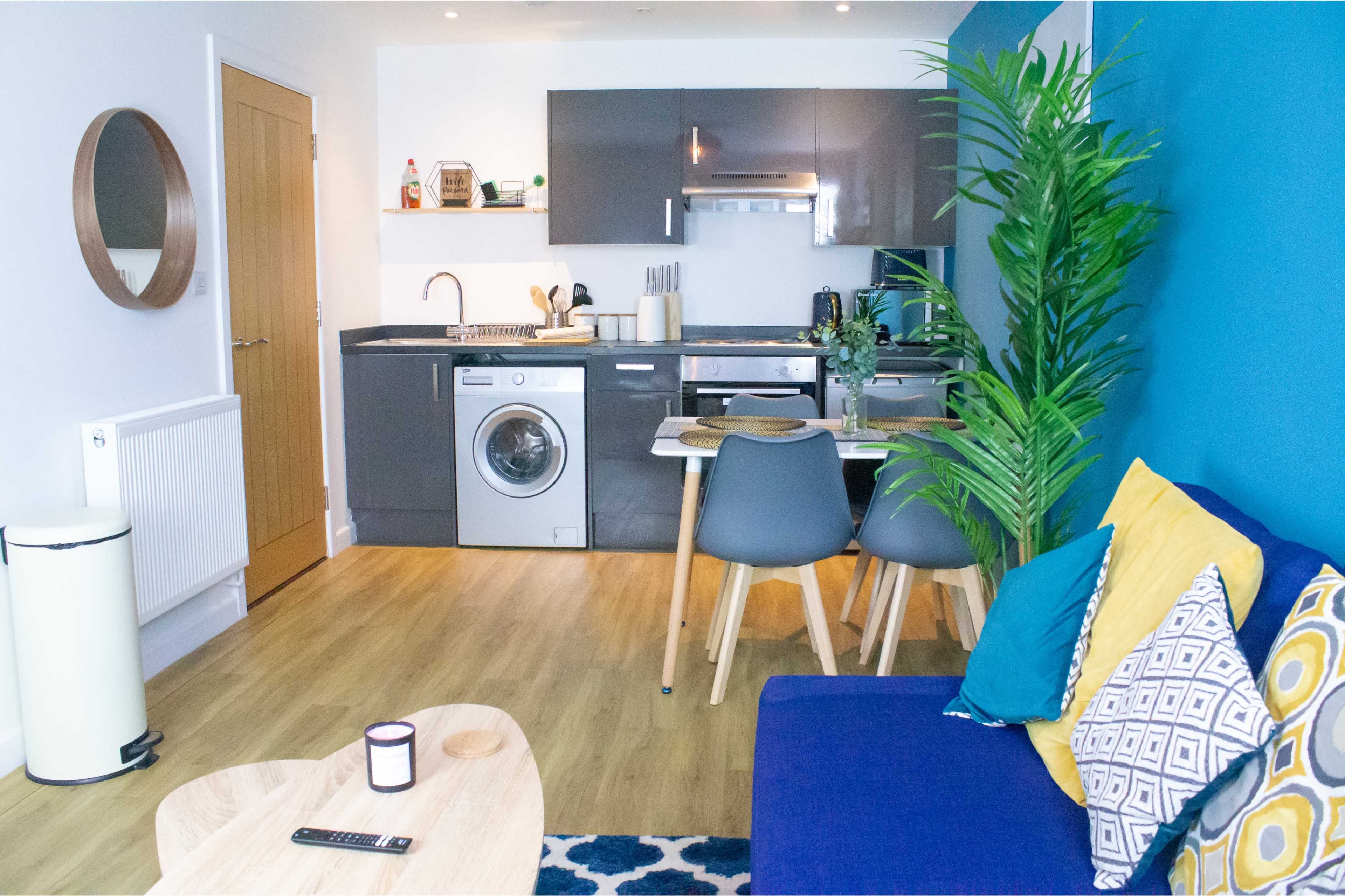A modern kitchen and living area with a washing machine, dining table, and colorful sofa against a blue wall.