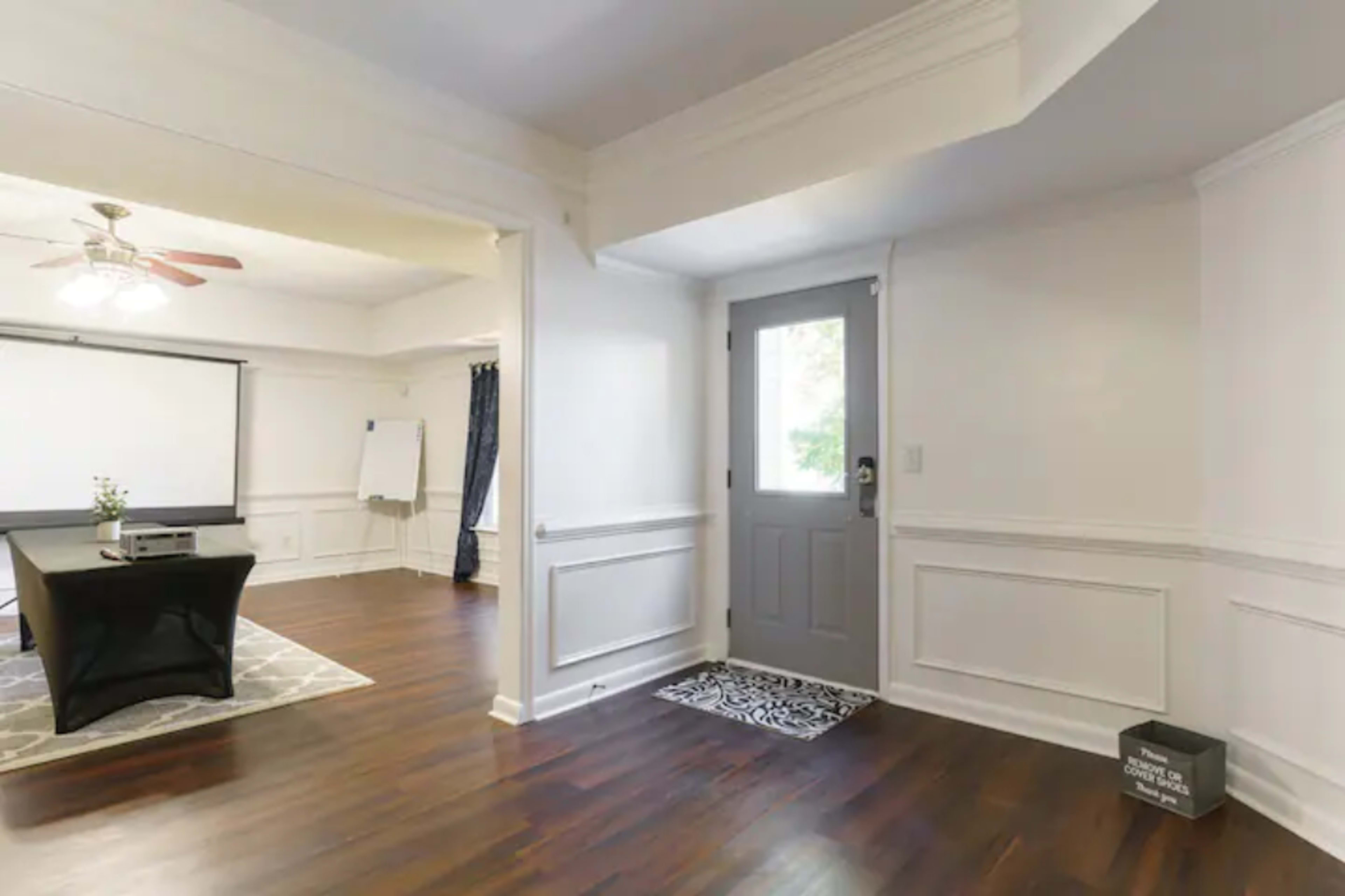 The image shows an interior space with a wooden floor, a door leading outside, and a small table in front of a projection screen.
