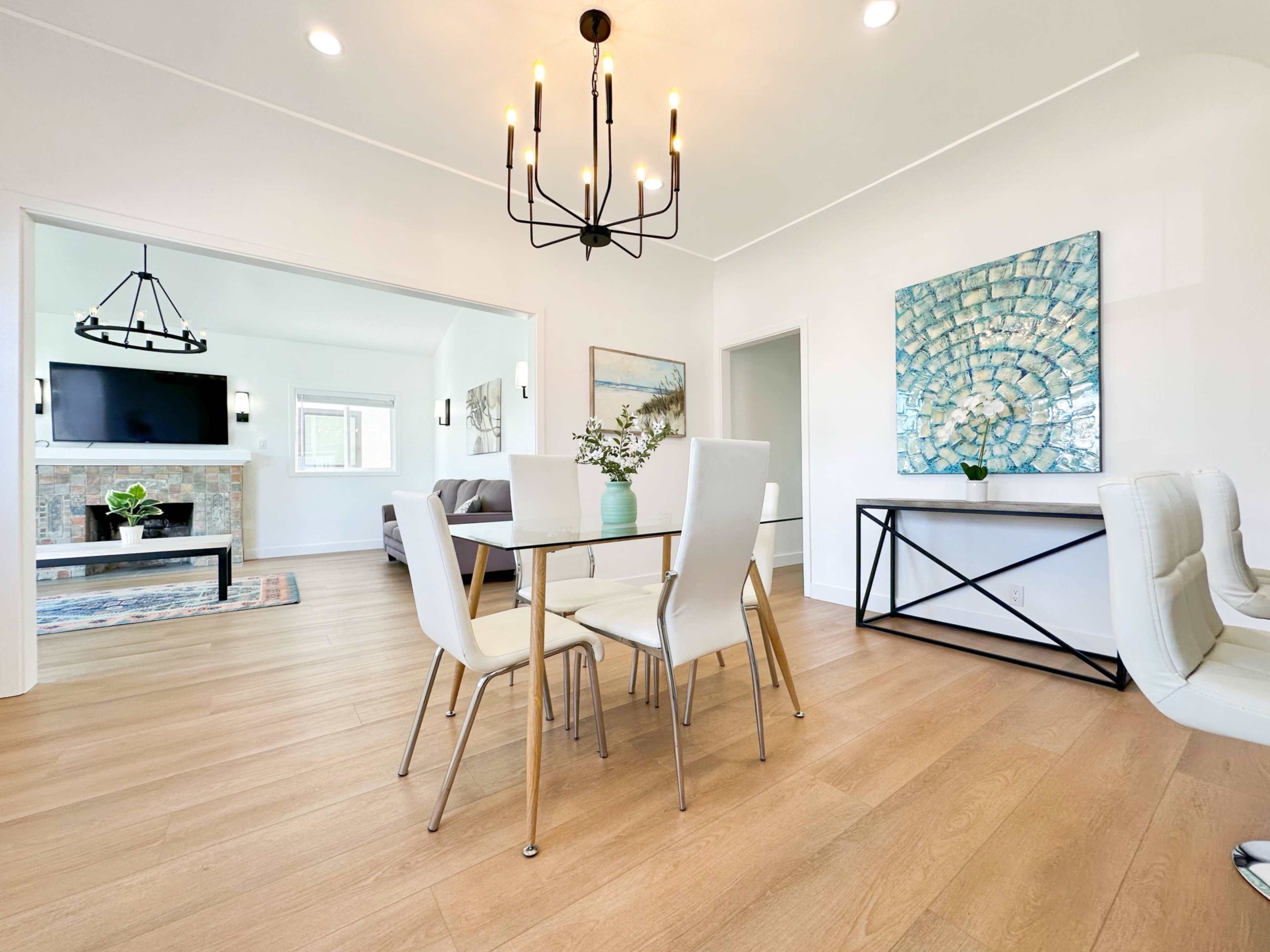 The image shows a modern dining area with a glass-top table and white chairs, featuring a chandelier and a decorative wall art piece.