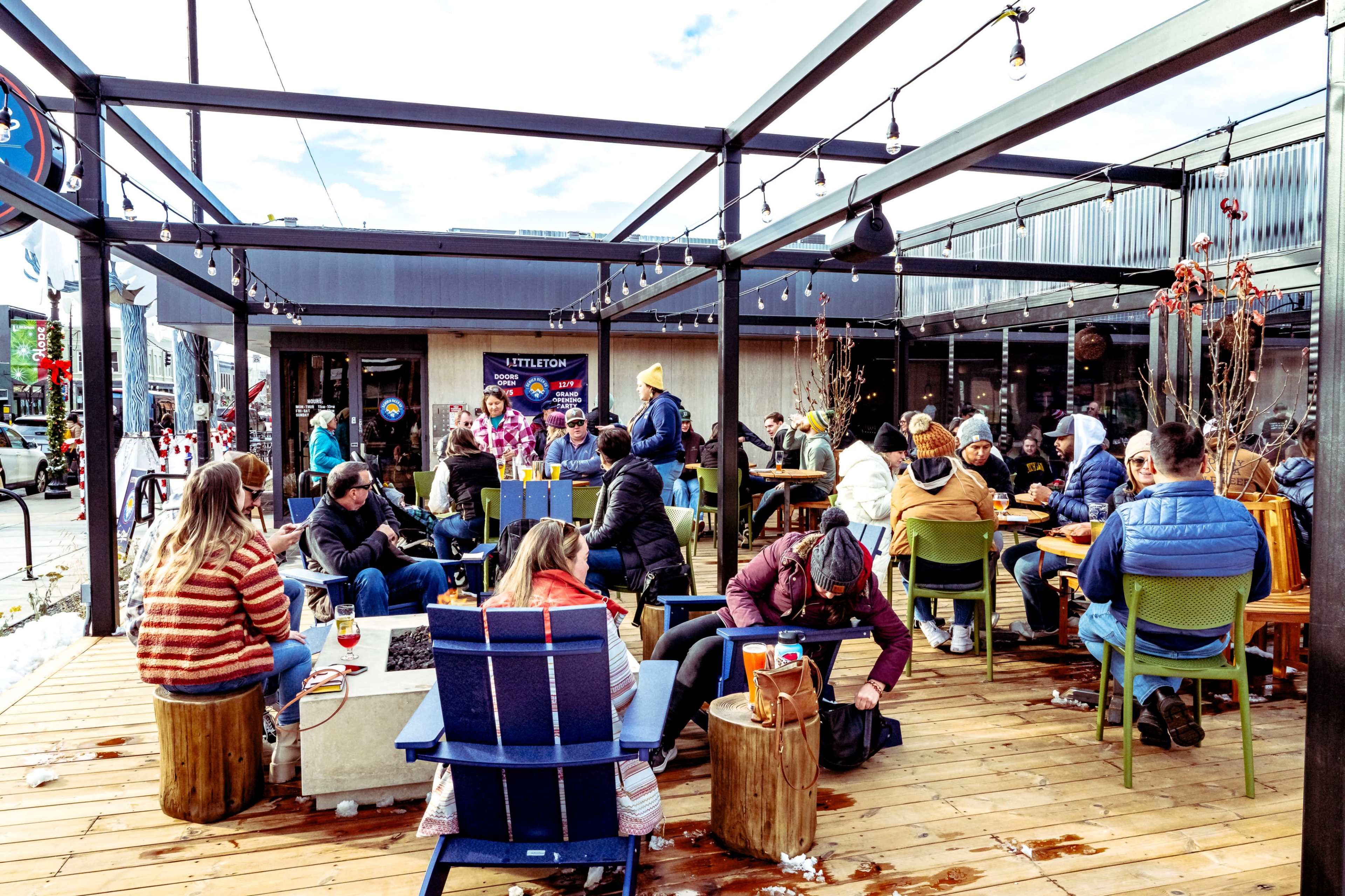 A lively outdoor seating area at a restaurant features groups of patrons enjoying food and drinks on wooden platforms under string lights.