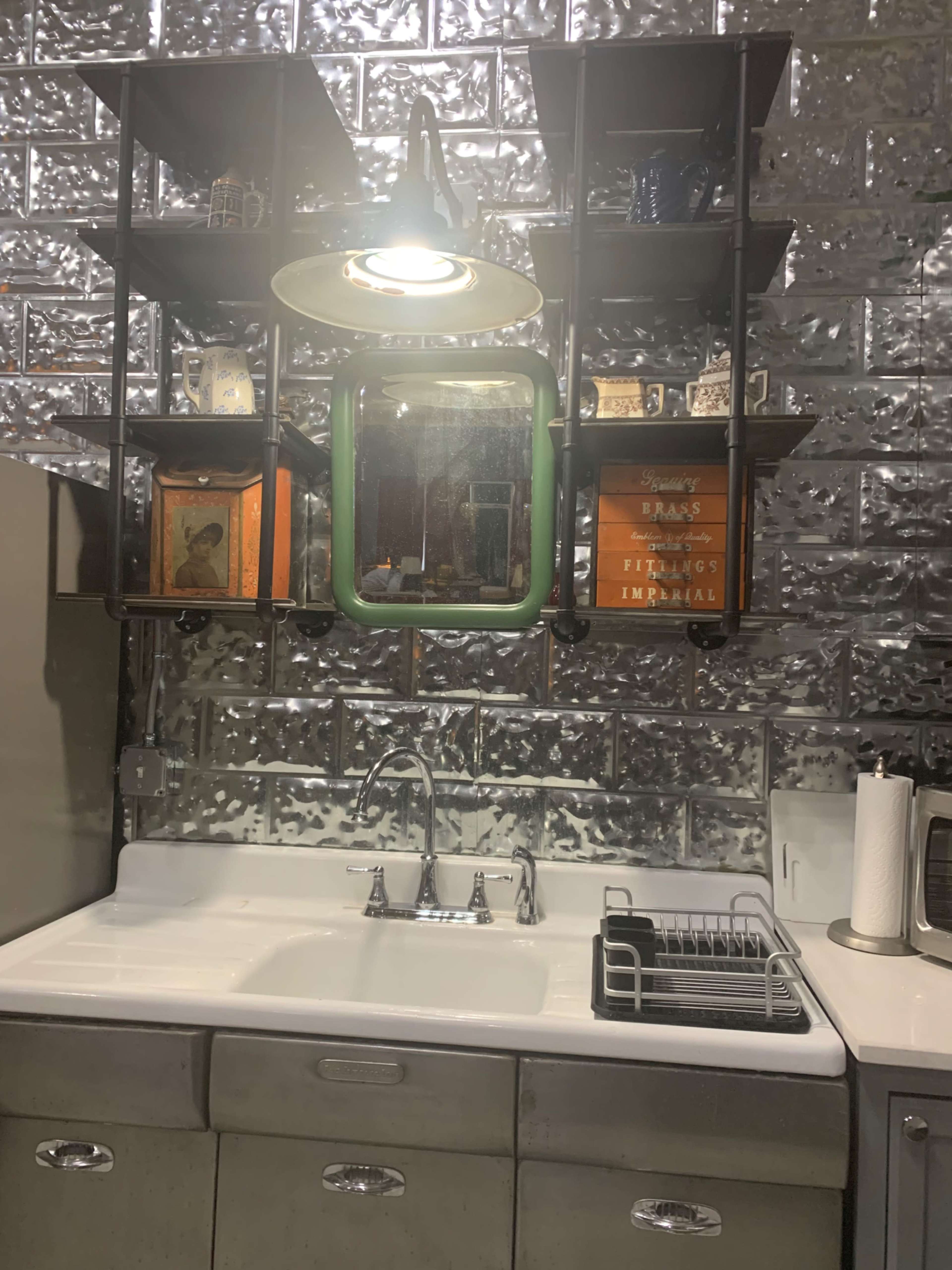 A modern kitchen with a stainless steel sink, shelving unit displaying various items, and a single overhead light.