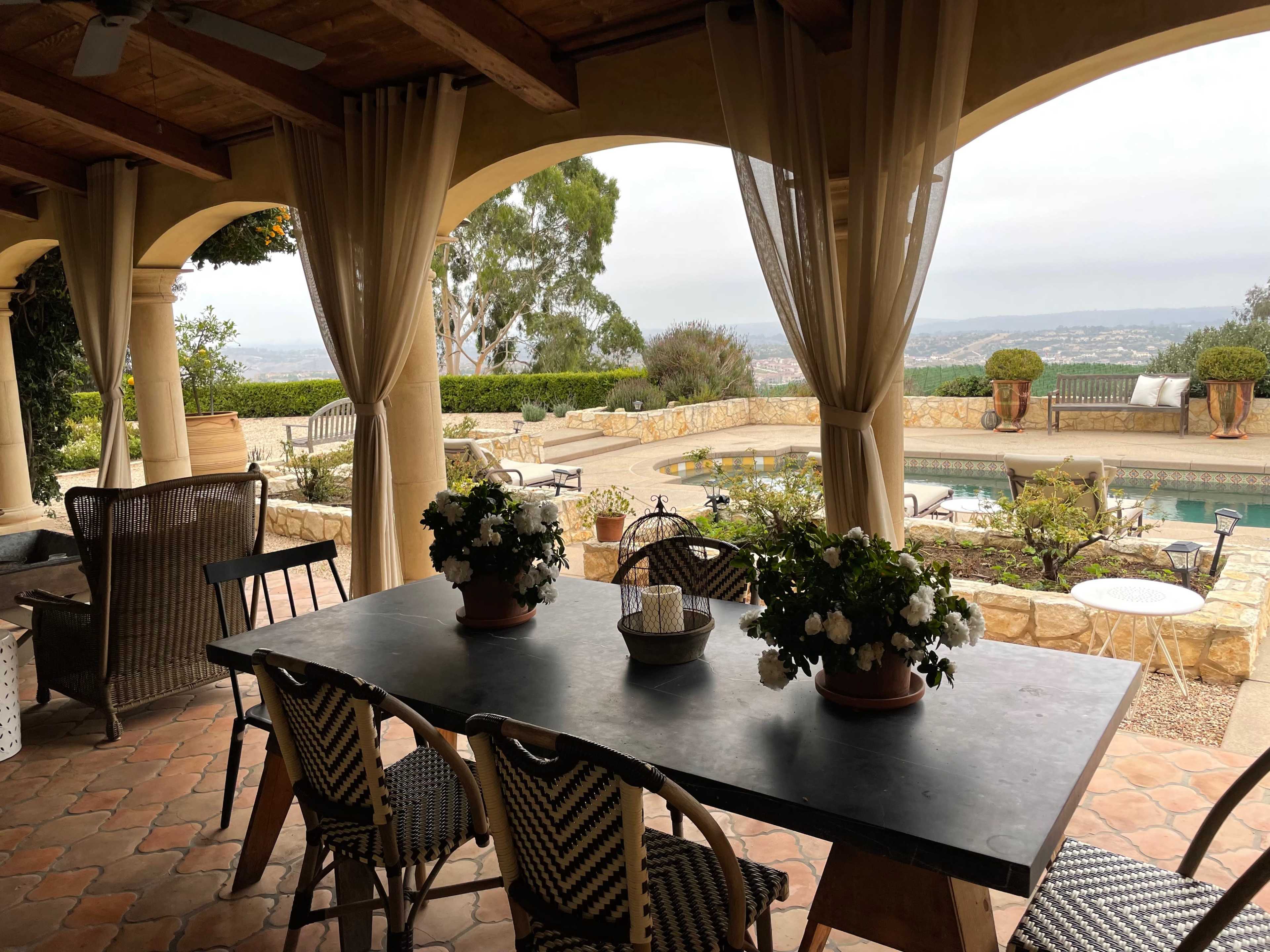 A covered outdoor dining area features a table with potted flowers, overlooking a pool and distant hills.