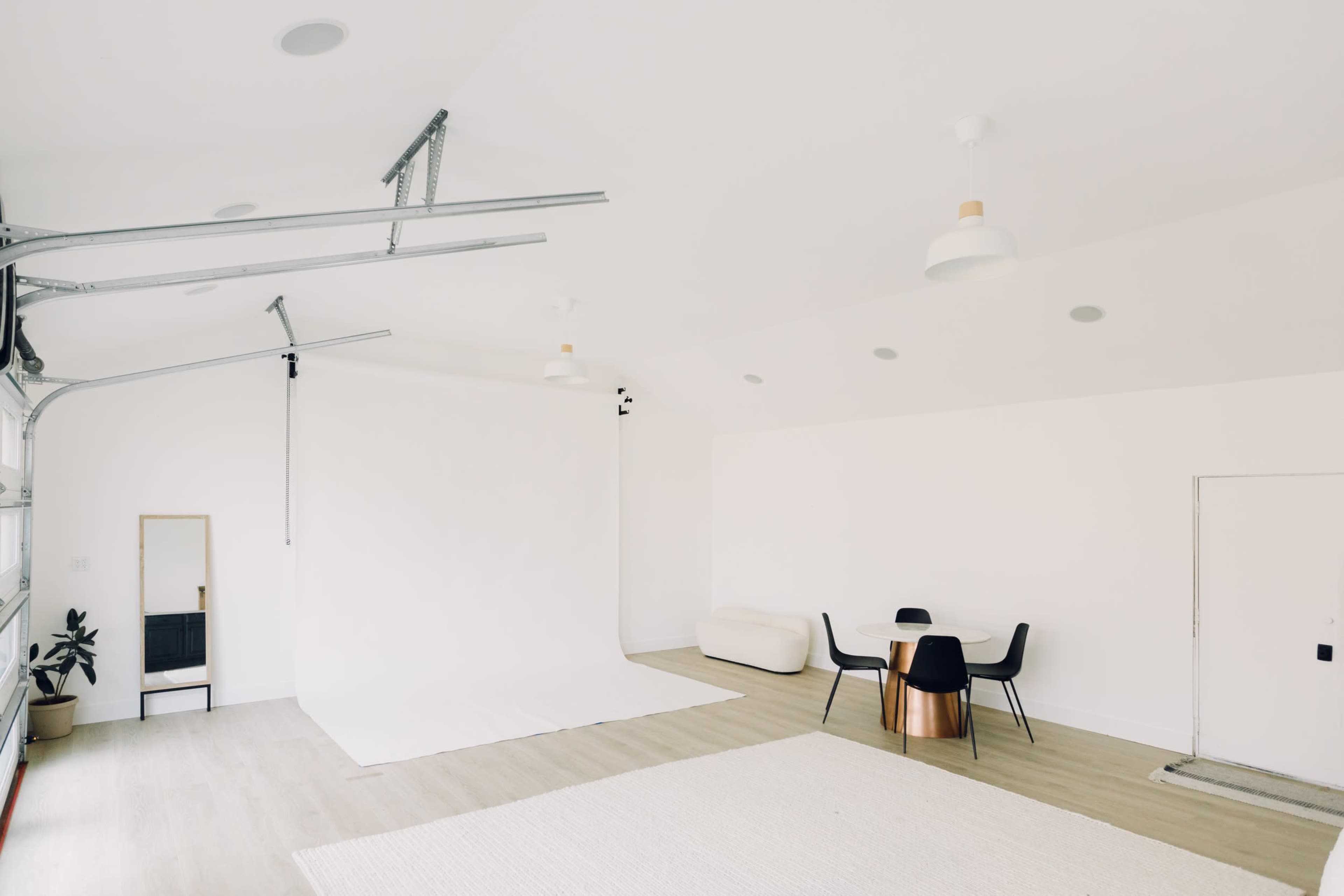 The image shows a minimalist studio space with a white backdrop, a round wooden table with black chairs, and a light-colored rug on the floor.