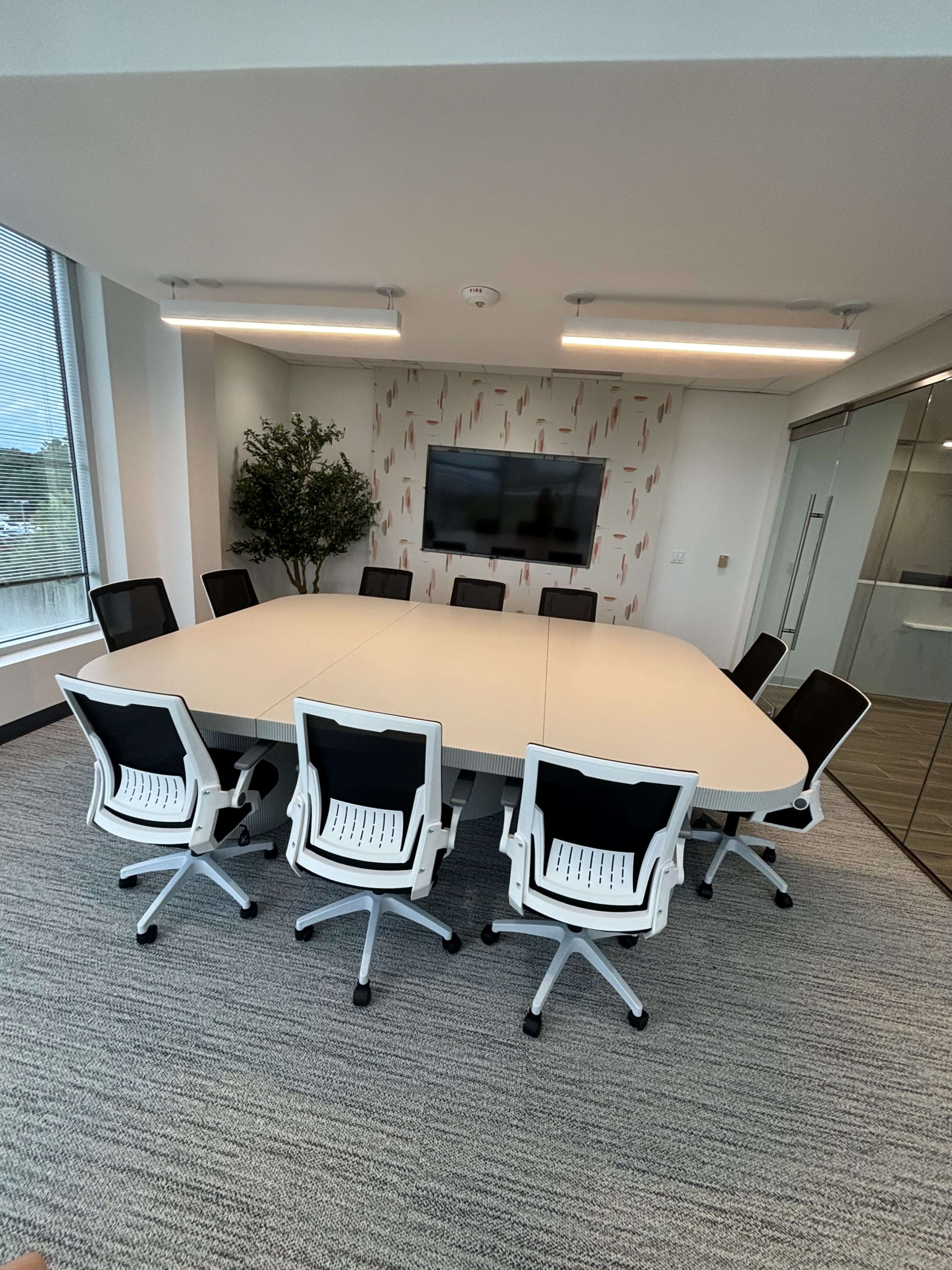 A modern conference room features a large oval table surrounded by ergonomic chairs, with a wall-mounted TV and large windows providing natural light.