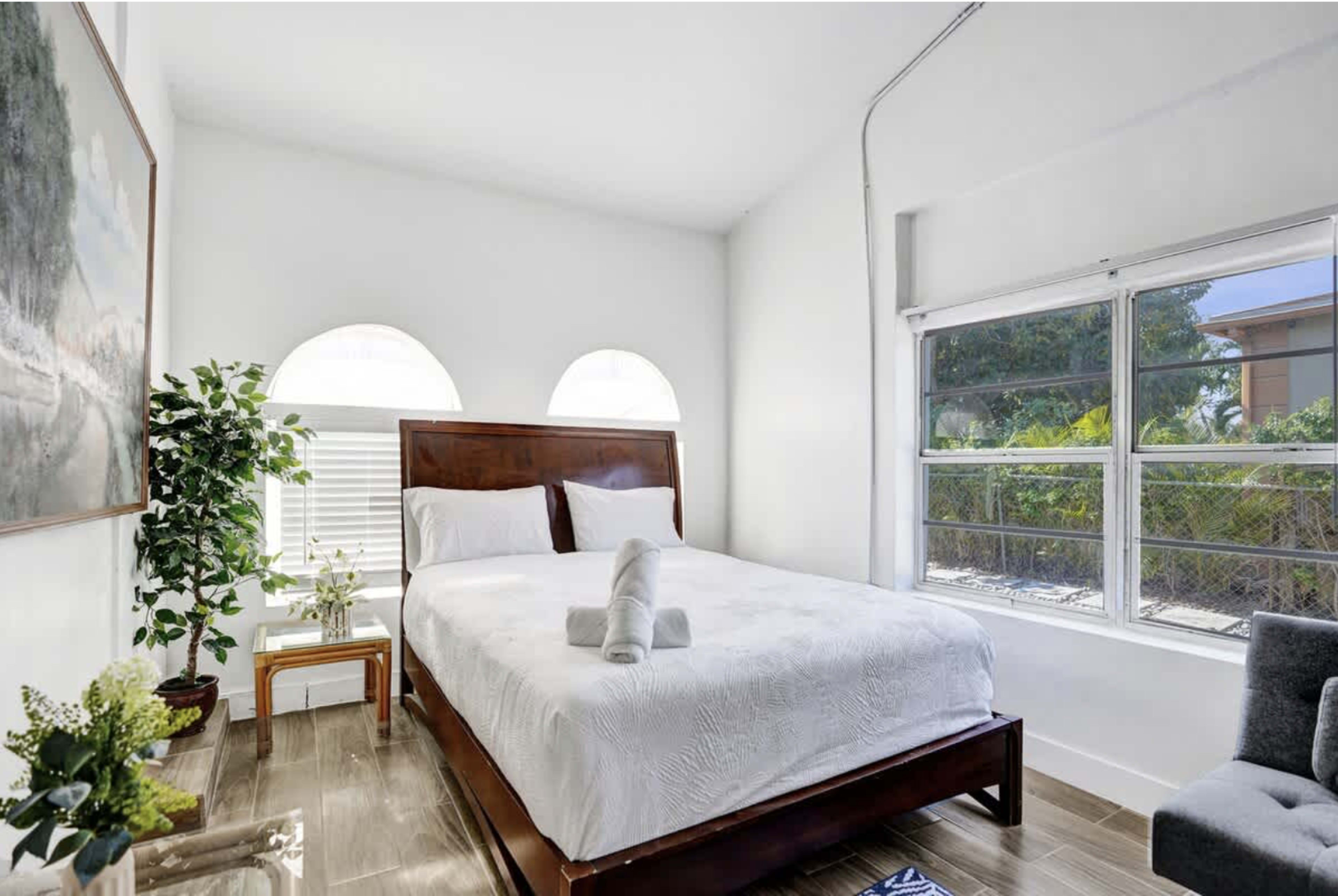 The image shows a well-lit bedroom featuring a queen-sized bed with white bedding, a small side table, and a potted plant near a window.