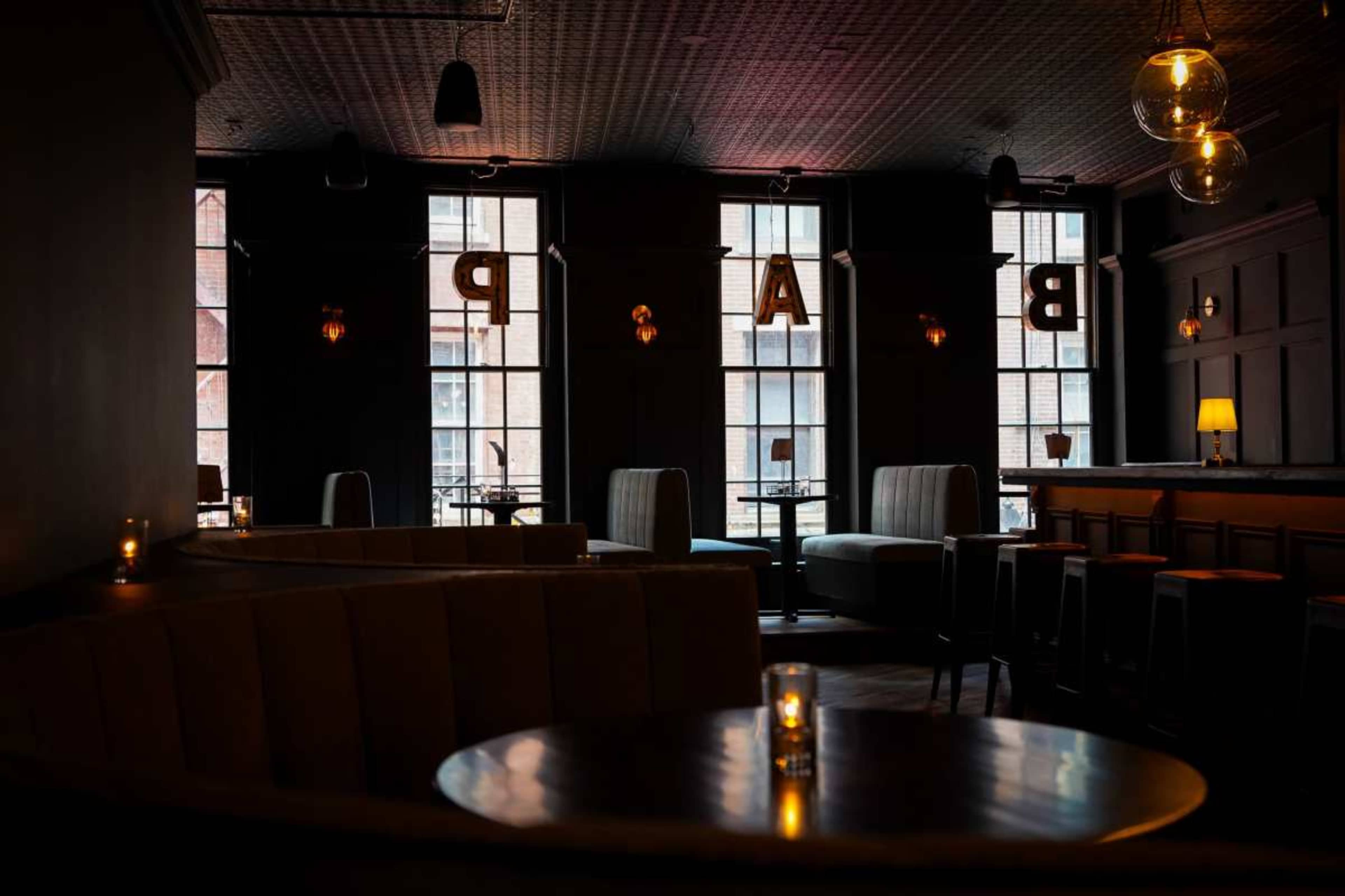 The image shows a dimly lit bar interior with a curved seating area, tables, and large windows featuring the word "BAR" illuminated.