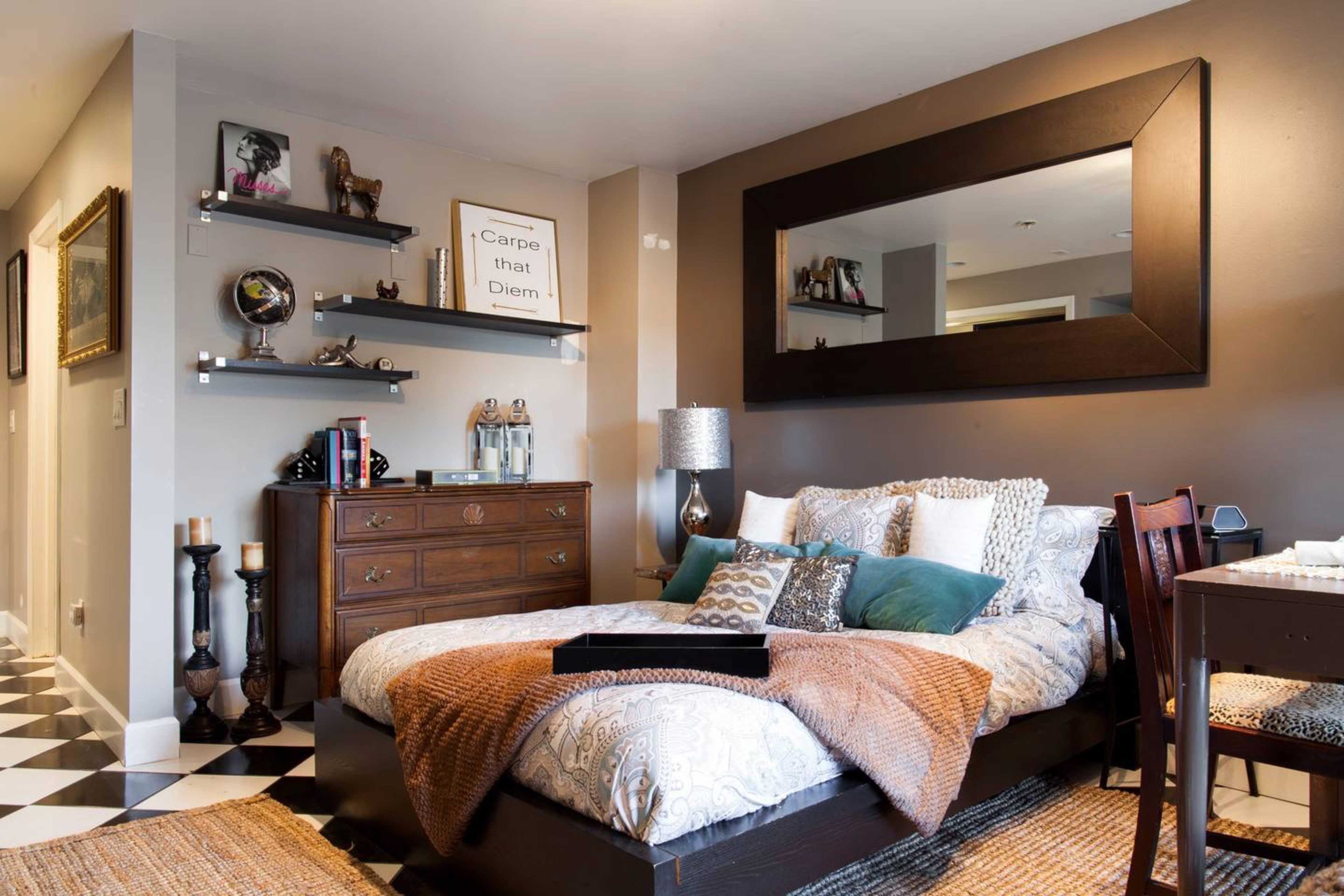A cozy bedroom with a large bed adorned with various pillows and textured throws, a wooden dresser, and shelves displaying decorative items on a checkered floor.