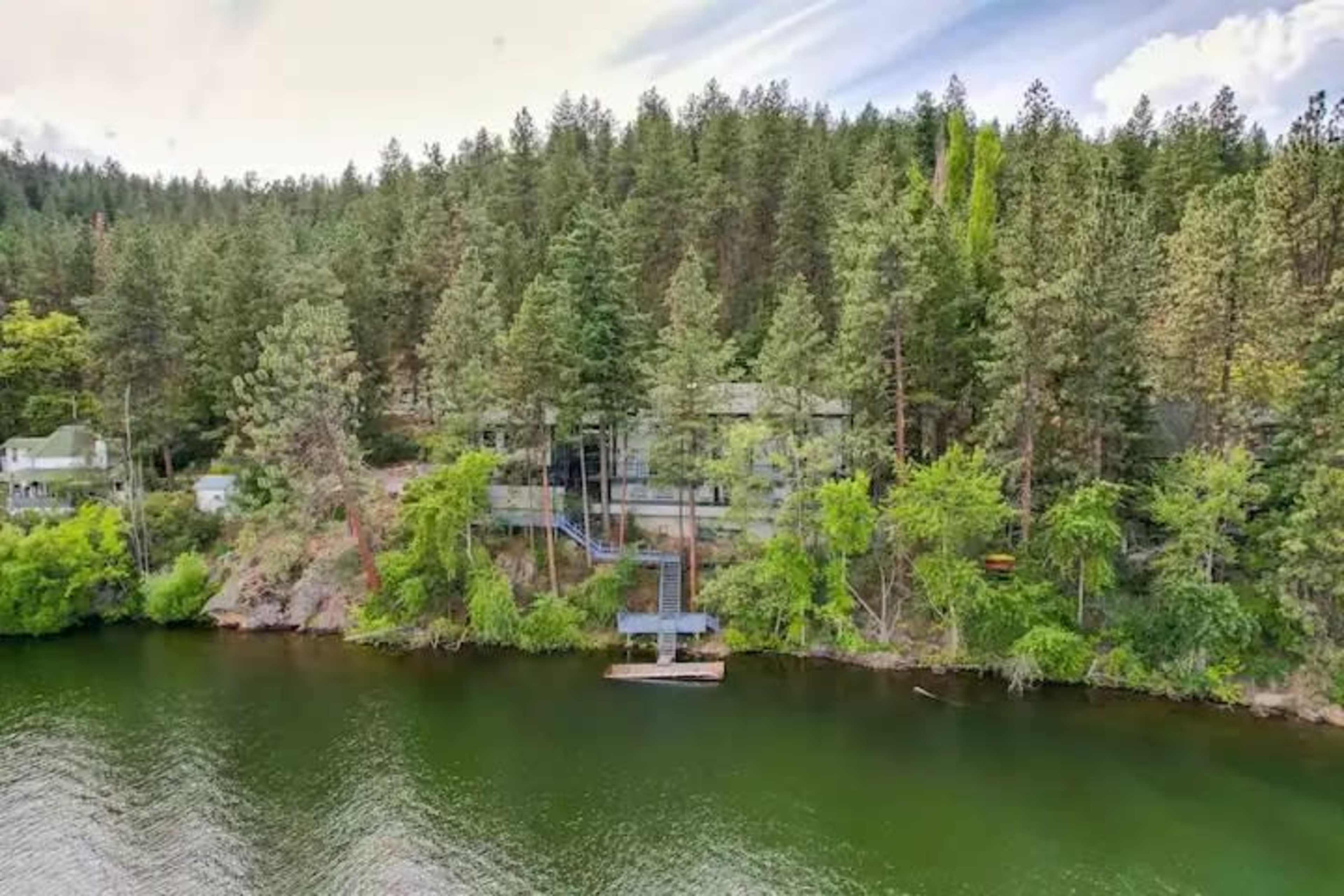 A modern house set among tall trees beside a calm lake, featuring a staircase leading down to the water.