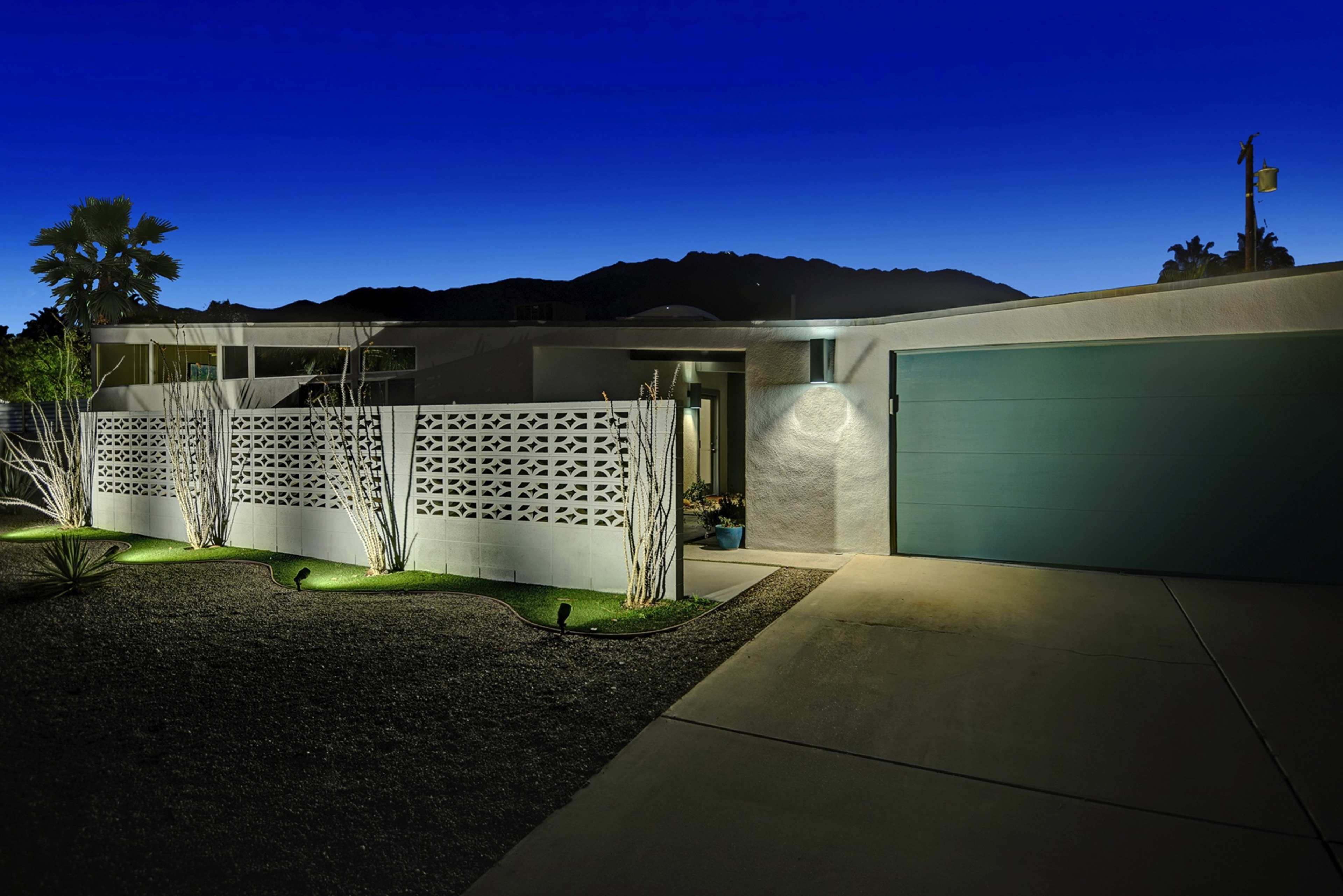 The image shows a modern house at dusk, featuring a decorative wall and a garage door, with landscaping illuminated by soft lights.