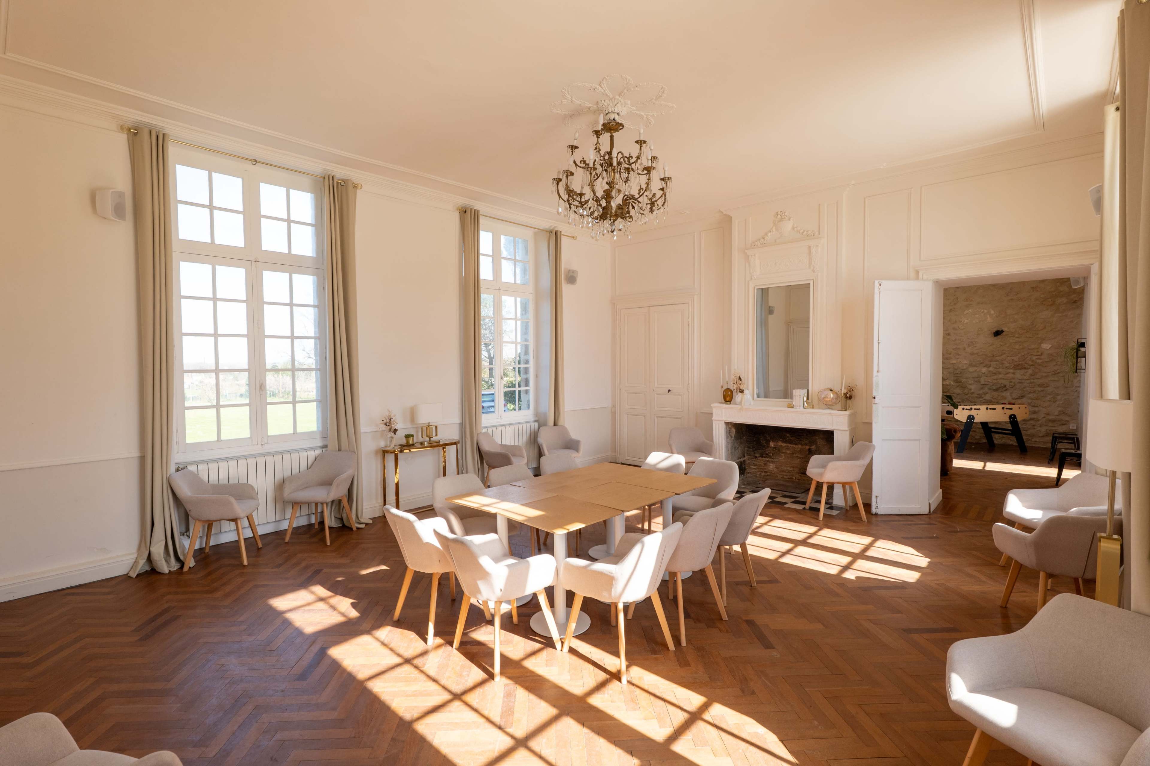 A spacious room features a wooden table surrounded by light-colored chairs, with large windows allowing natural light to illuminate the space.
