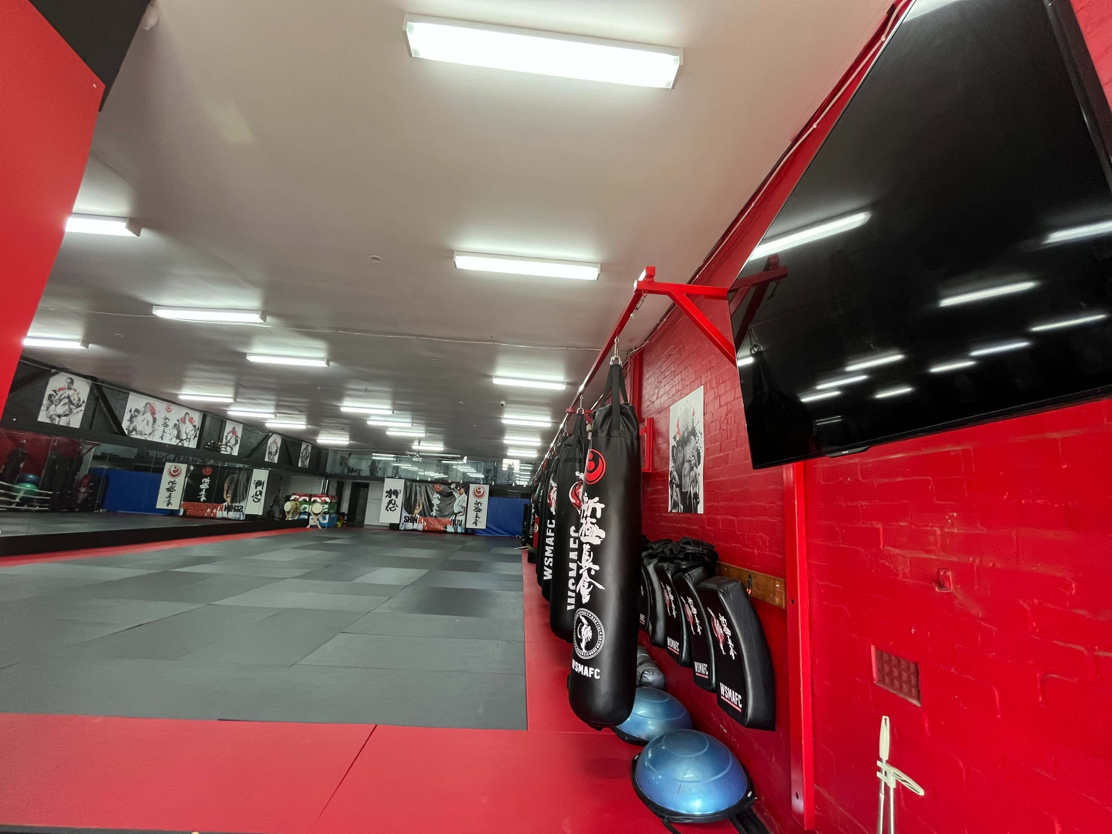 The image shows an empty martial arts studio featuring hanging punching bags, exercise balls, and wall-mounted television screens, with a mirrored wall in the background.