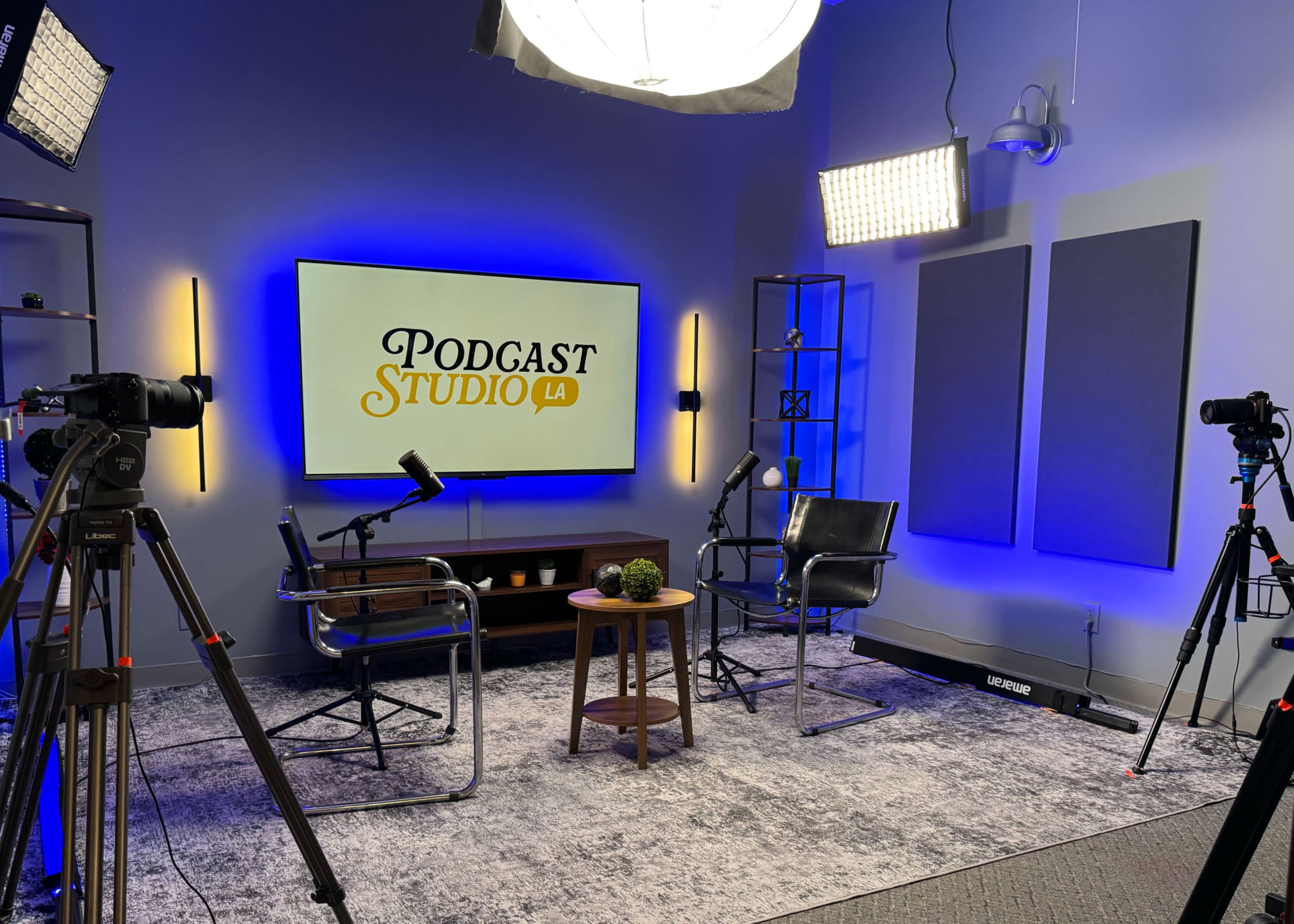 The image shows a podcast studio with two black chairs, a small wooden table, and professional lighting set up in front of a large screen displaying "PODCAST STUDIO LA."
