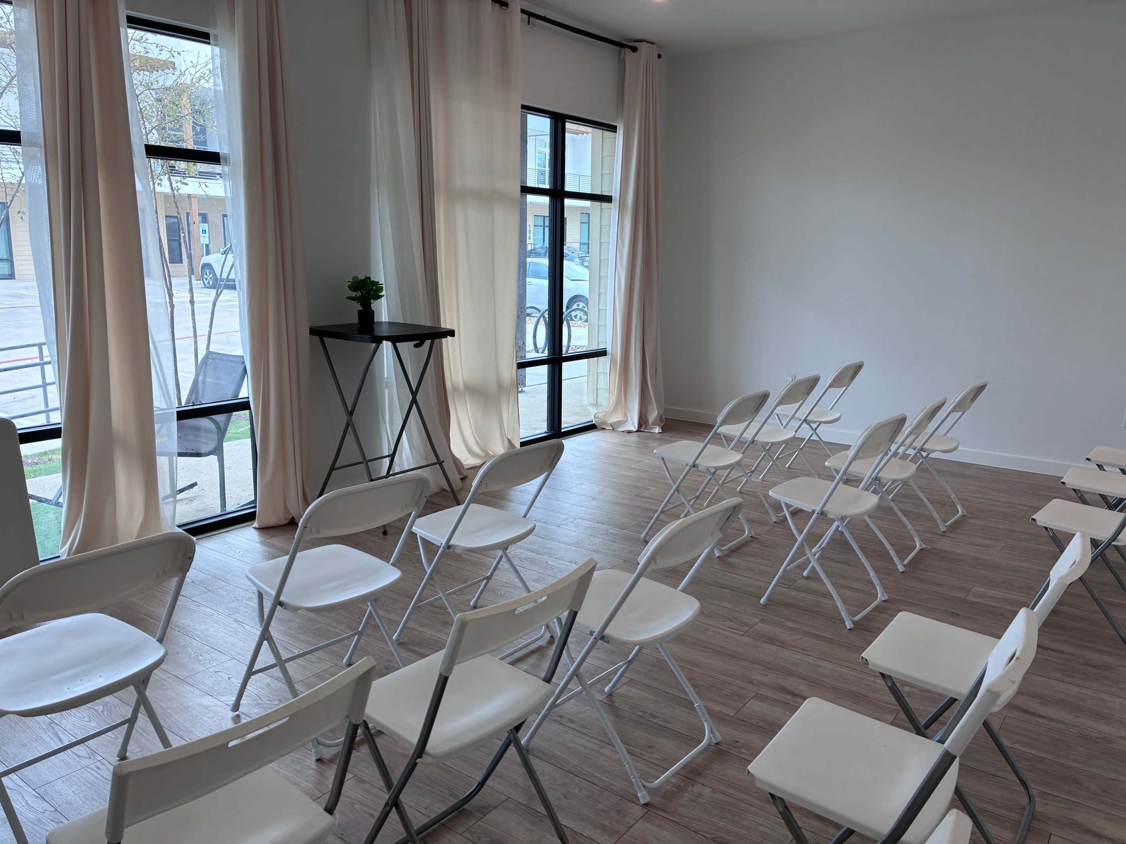 The image shows a minimalistic room with rows of white folding chairs facing a small black table beside large windows with light curtains.